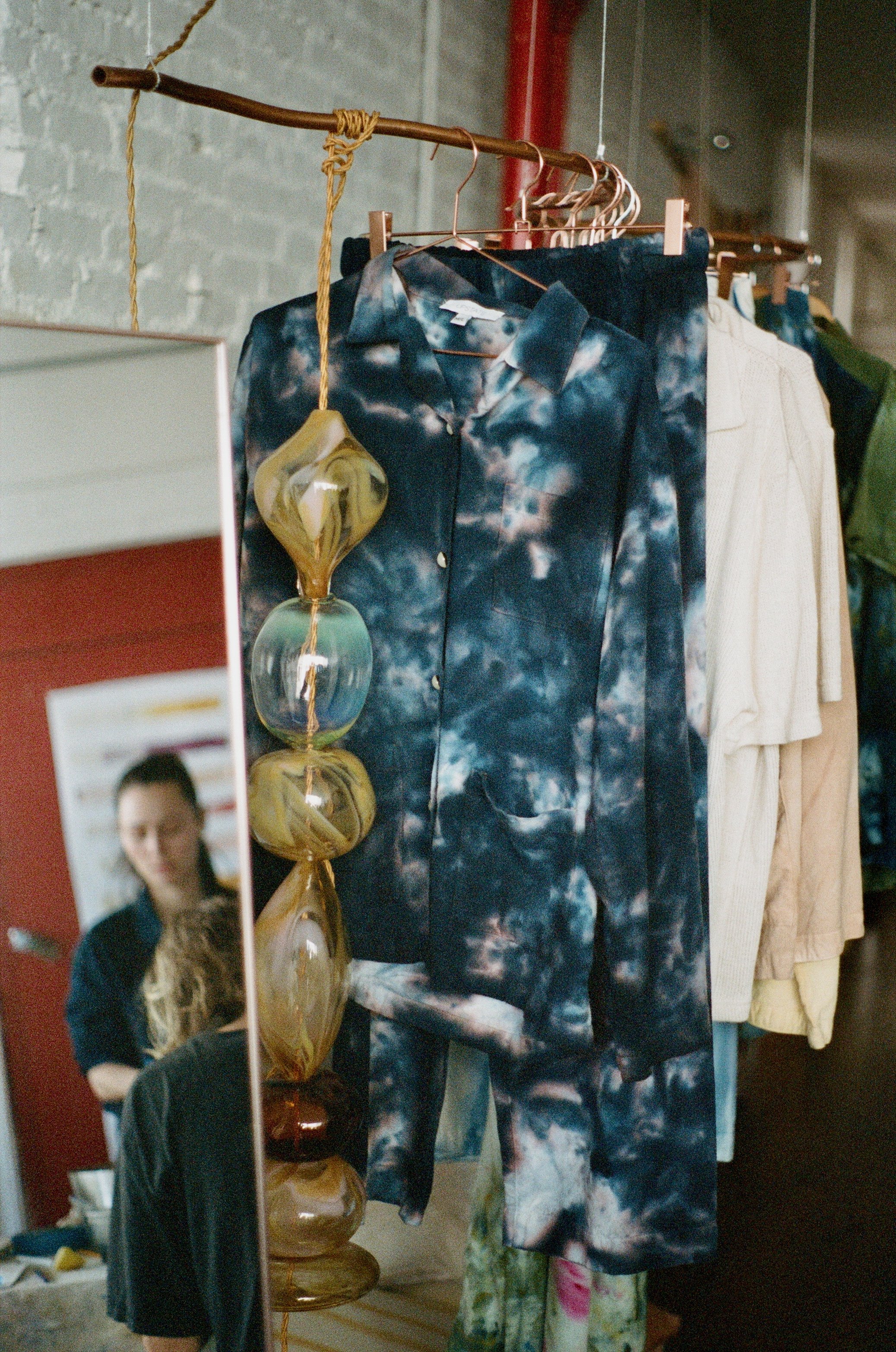 Clothing rack with tie-dye shirt and other garments, next to a mirror reflecting a woman and a mannequin.