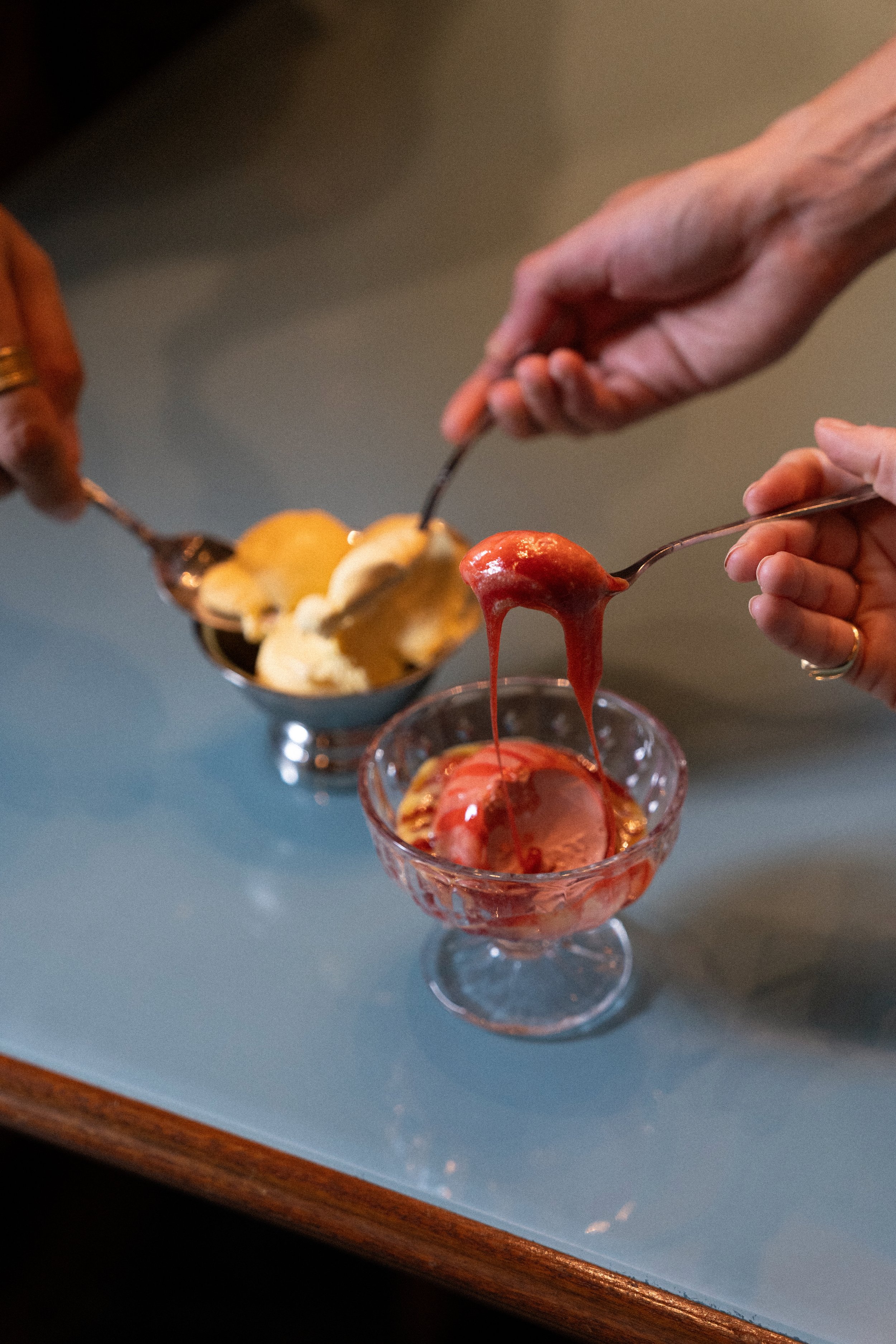 People scooping ice cream into a glass bowl, with the ice cream melting and drizzling down.