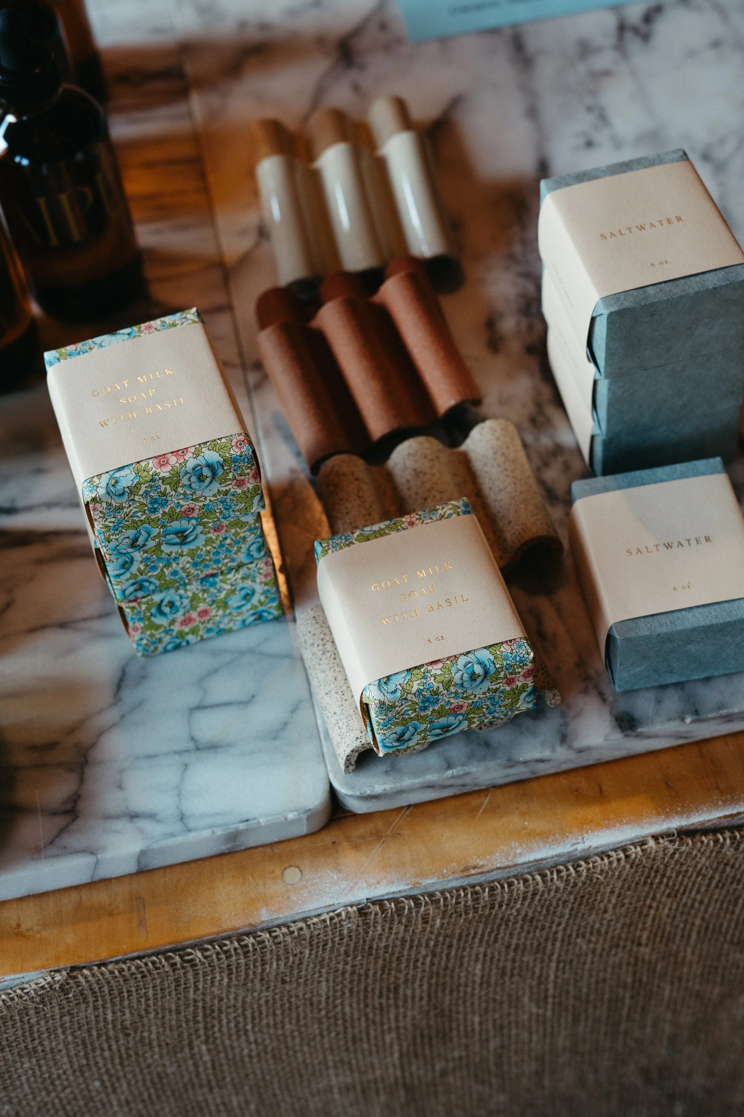 Various skincare products on marble and wooden surfaces, including goat milk soap with basil, saltwater soap, and small bottles with wooden caps.