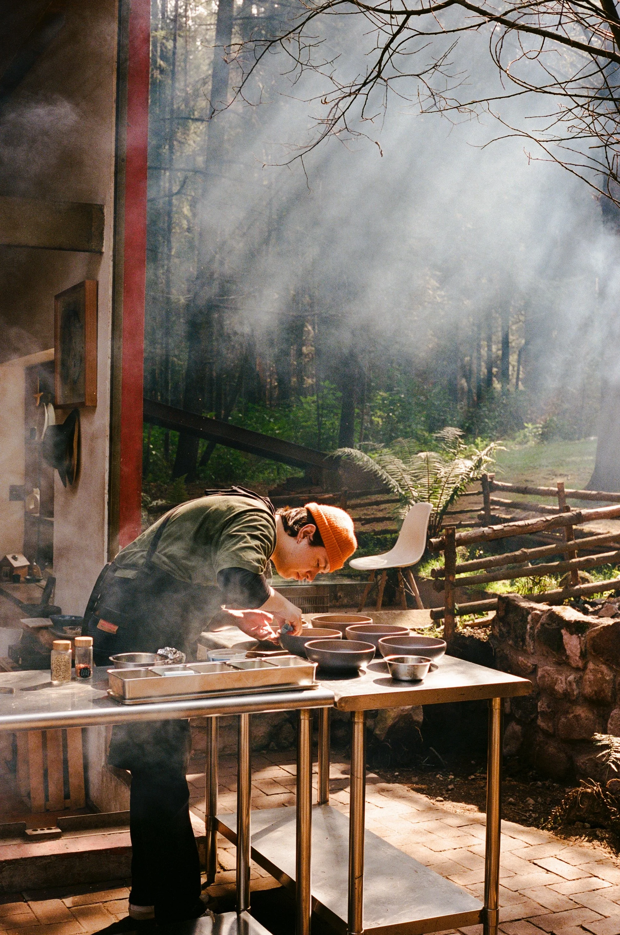 A person wearing an orange beanie and dark clothing preparing food on a stainless steel outdoor table, with a forest scene backdrop and sunlight filtering through trees.