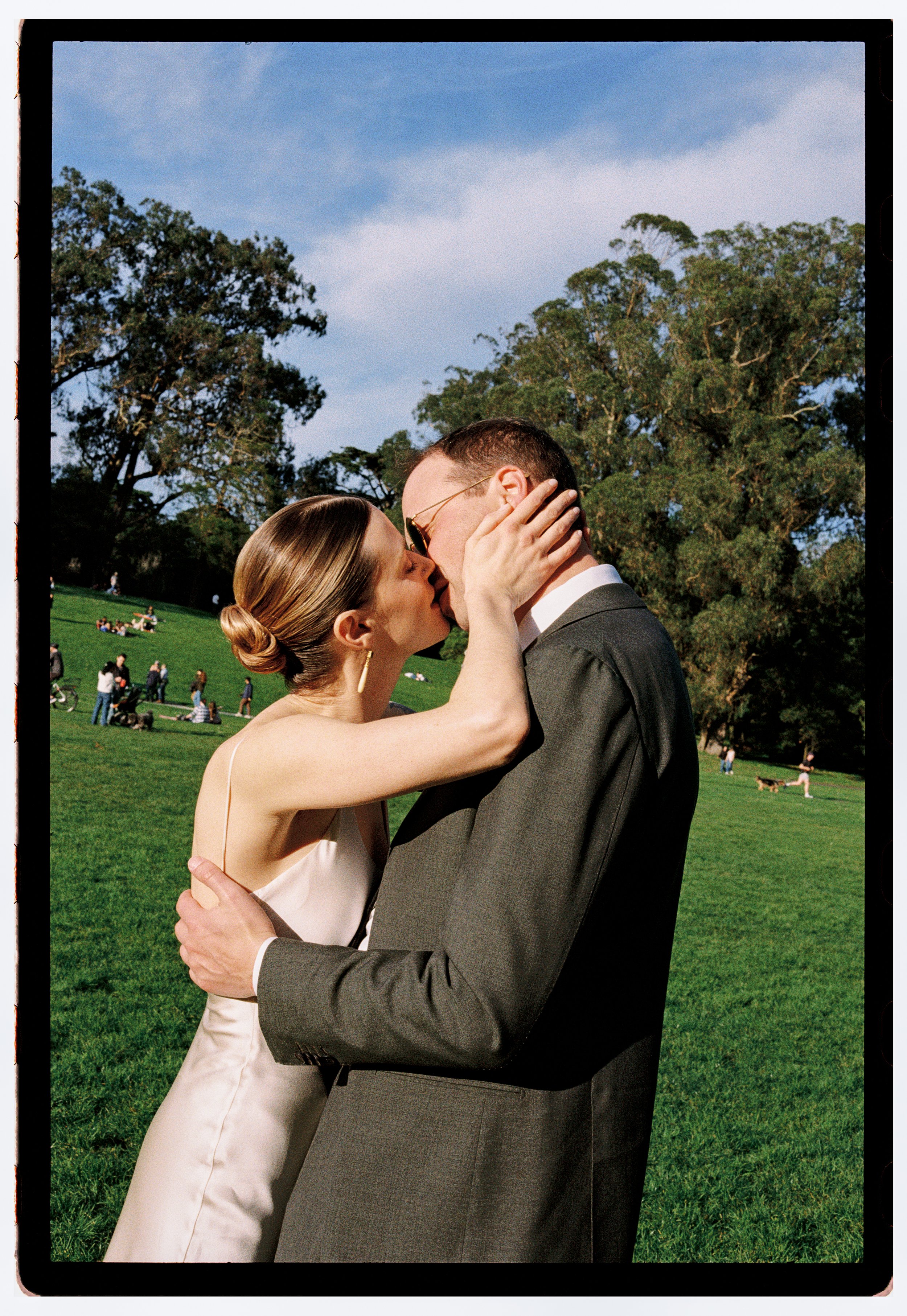 A couple sharing a kiss in Golden Gate Park with green grass and trees, people lounging, and dogs in the background on a sunny day.