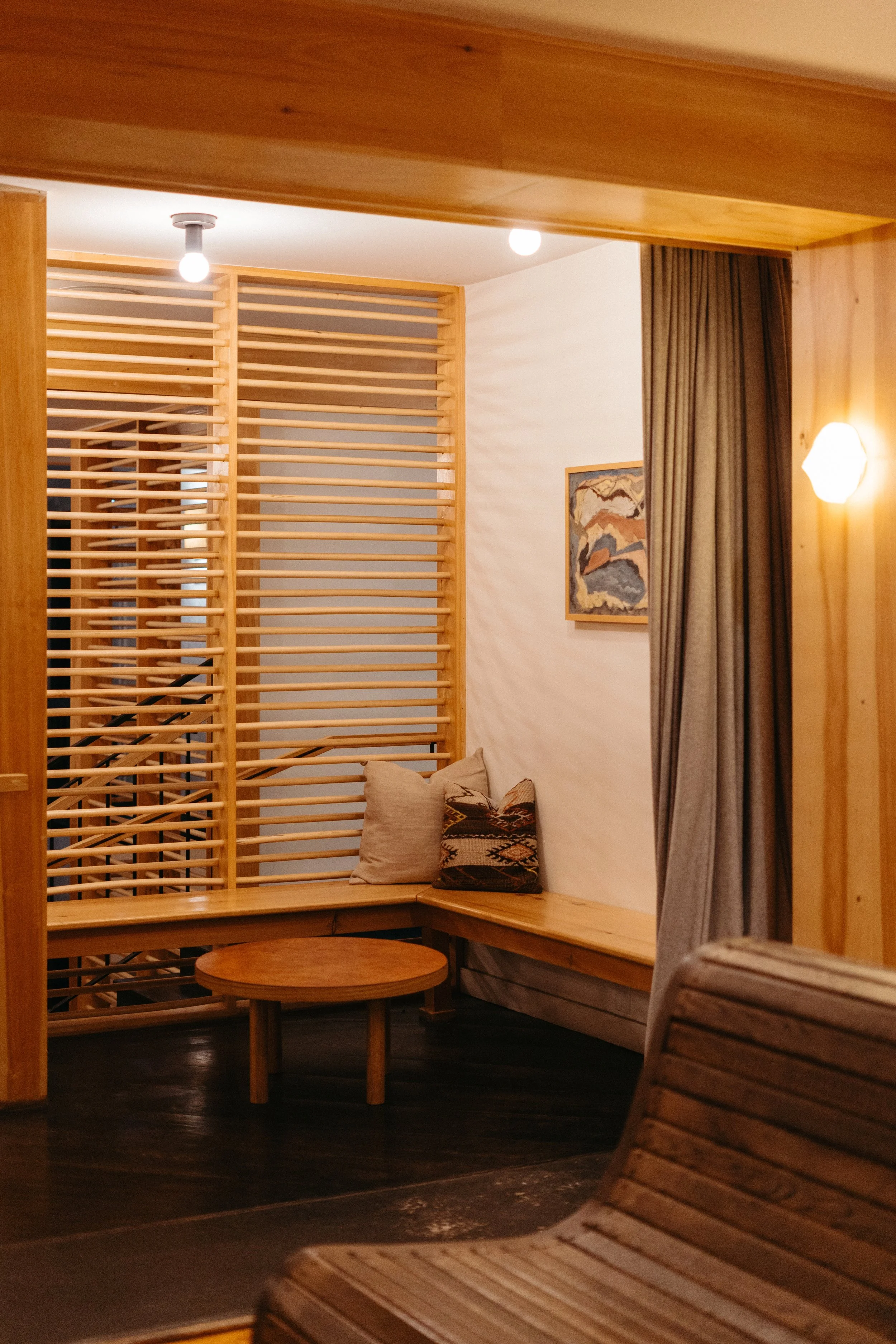 Cozy interior space with wooden furniture, a small round table, a built-in wooden bench with pillows, a painting on the wall, a curtain, and warm lighting.