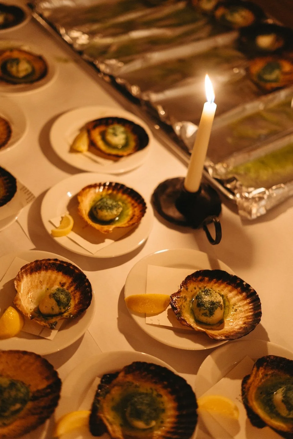 Several plates of baked scallops garnished with herbs and lemon wedges on a white table, lit by a single candle.