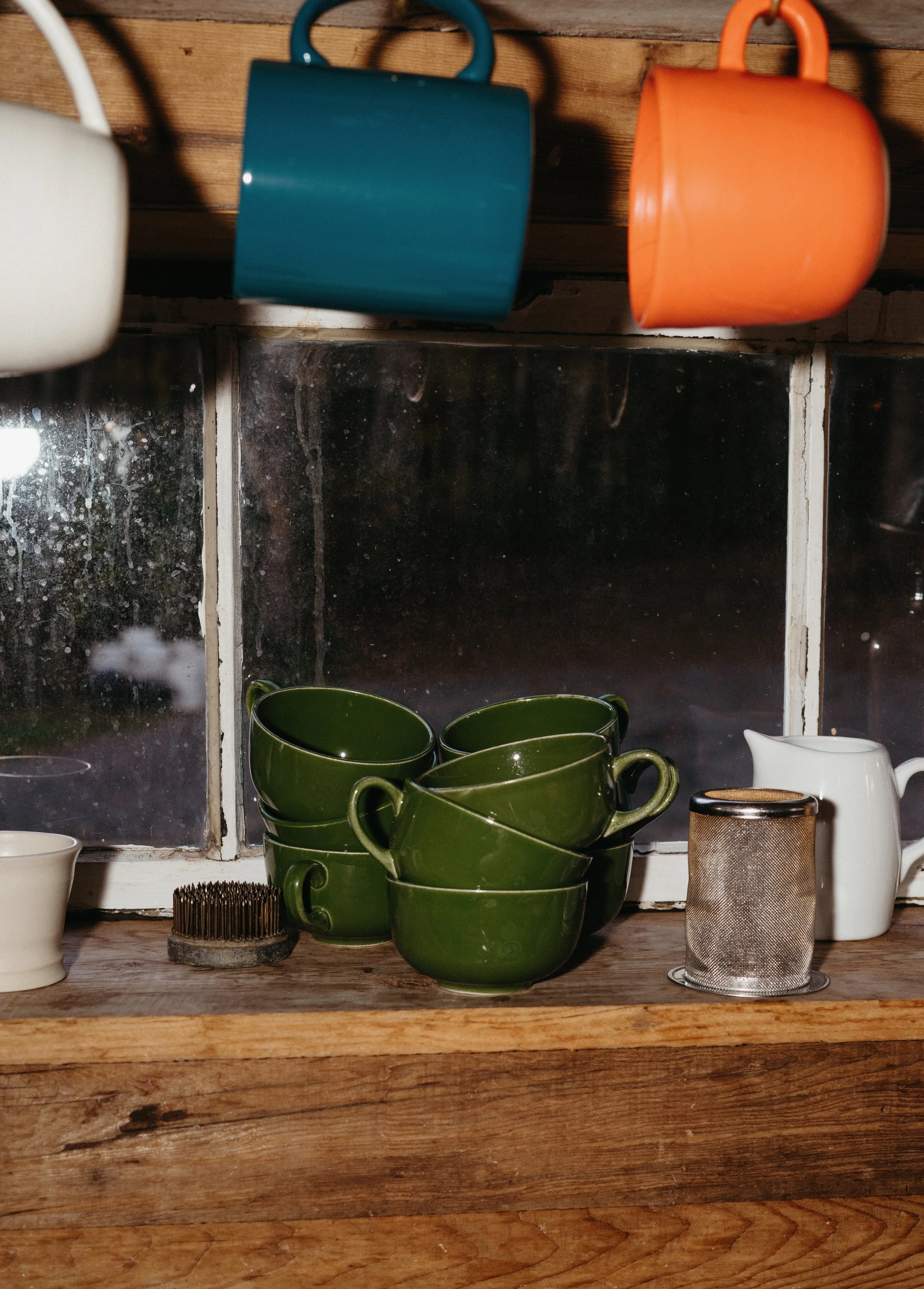 A rustic kitchen window sill with green coffee mugs stacked, a white creamer pitcher, a metal tea strainer, a small white bowl, and a used coffee filter. Above, colorful mugs hang from hooks on a wooden board.