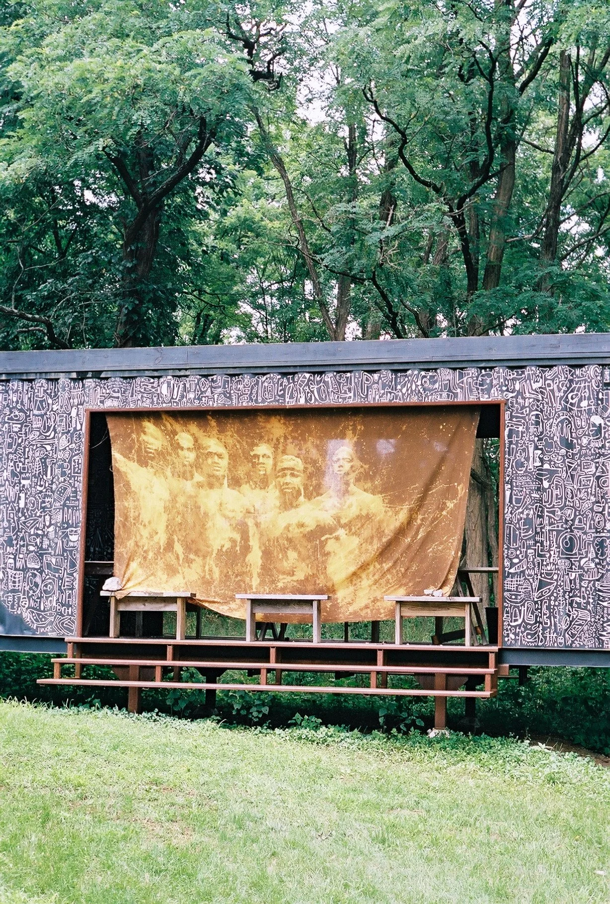 A stage with a backdrop of a yellow-brown historic photograph of several people, surrounded by a black frame decorated with white doodle artwork, located outdoors among green trees.
