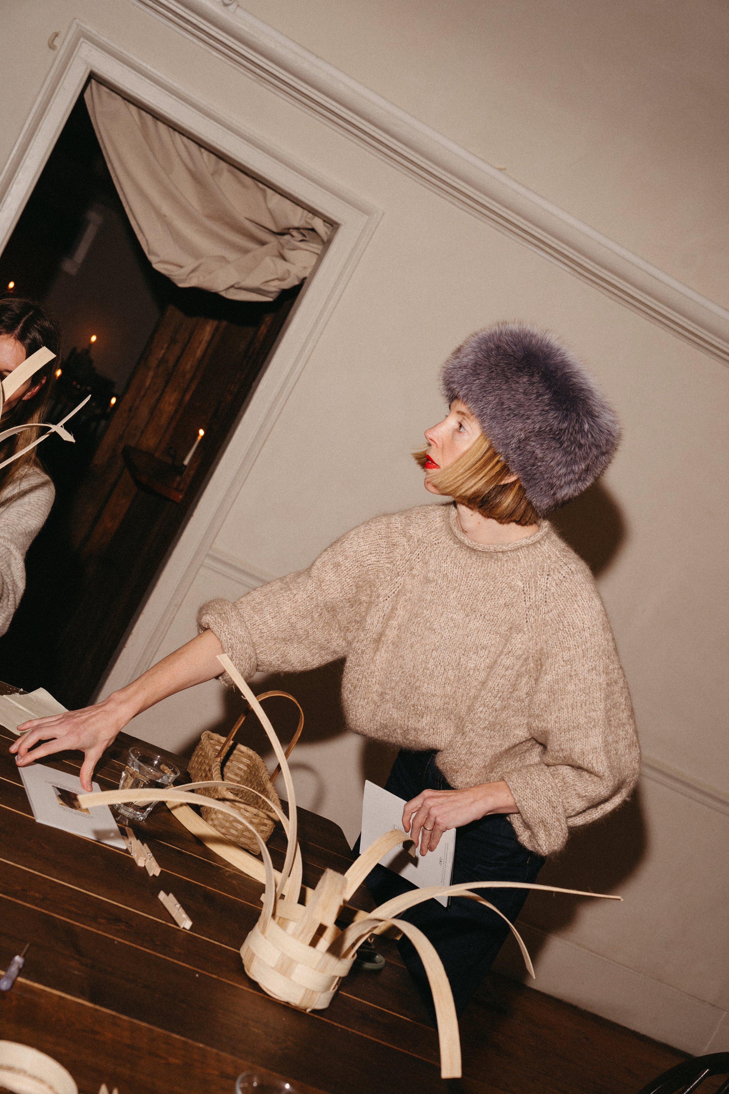 A woman wearing a fuzzy gray hat and a beige sweater reaching across a table with various items including a woven basket, paper, and a glass, with a window in the background showing candles outside.