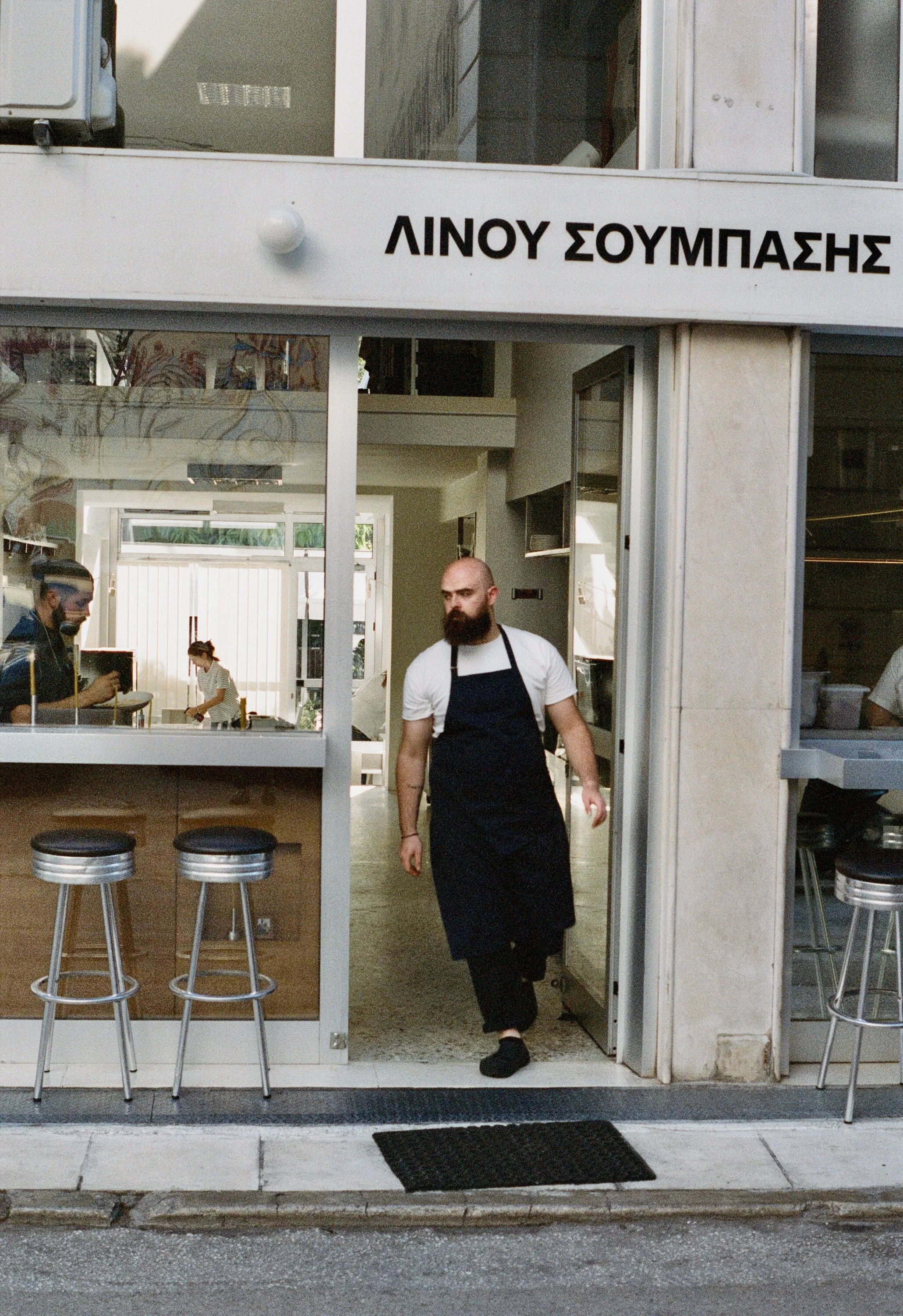 A man with a beard and wearing a white shirt and black apron walking out of a restaurant. The restaurant has Greek signage above the door, and there are bar stools outside. Inside, there are people preparing food.
