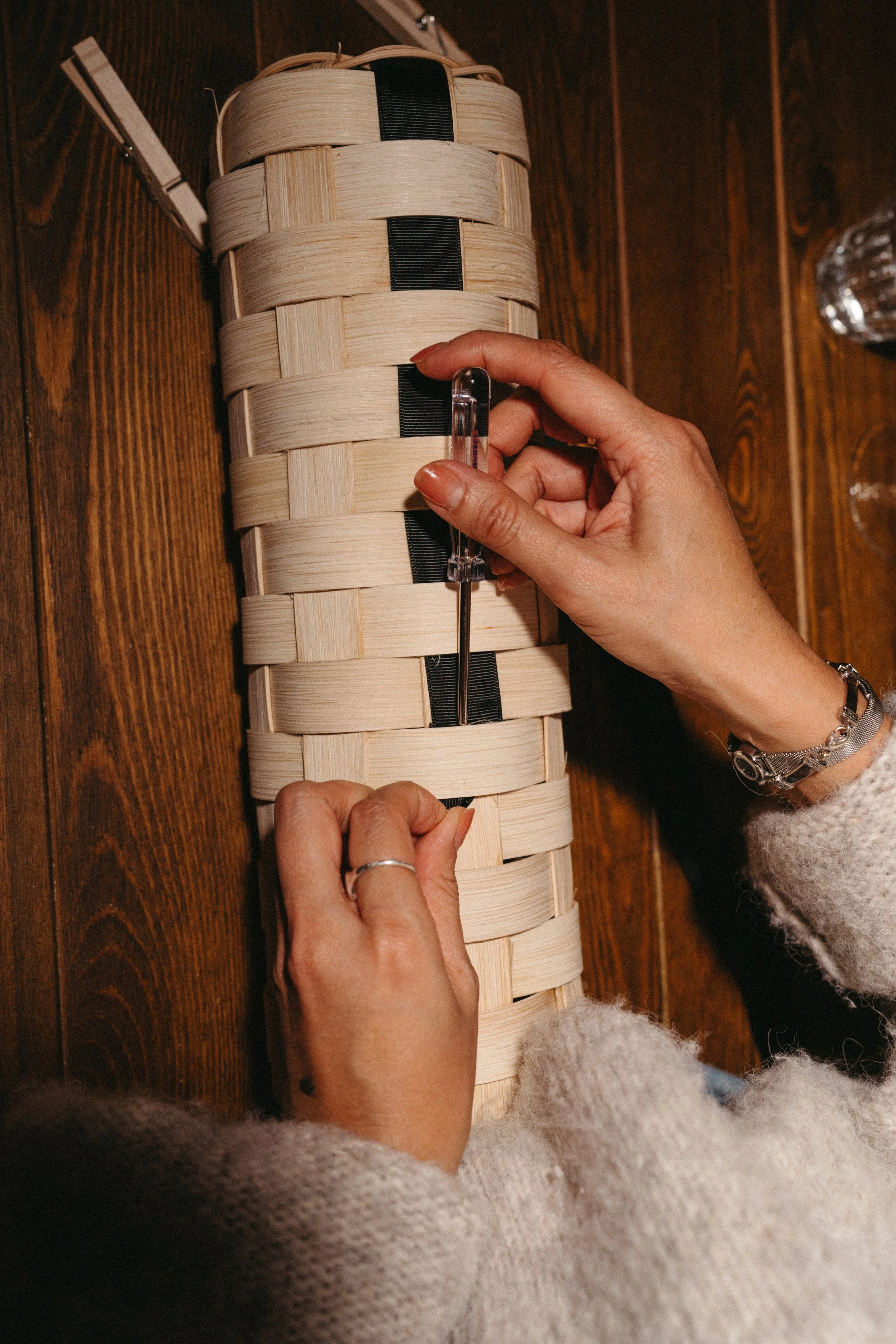 A person is using a screwdriver to work on a woven rectangular wooden object, possibly a basket, on a wooden surface.