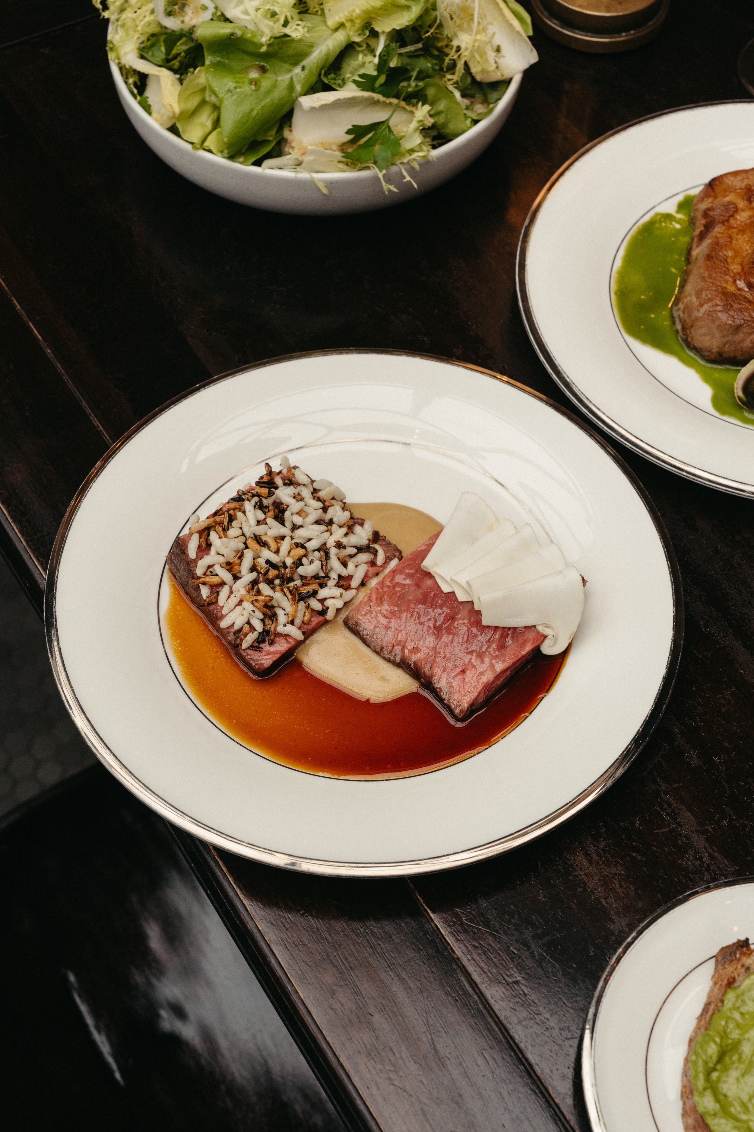 Plate of seared tuna topped with chopped nuts, served with a creamy sauce and sliced cheese, surrounded by other plates and a bowl of salad on a dark wooden table.