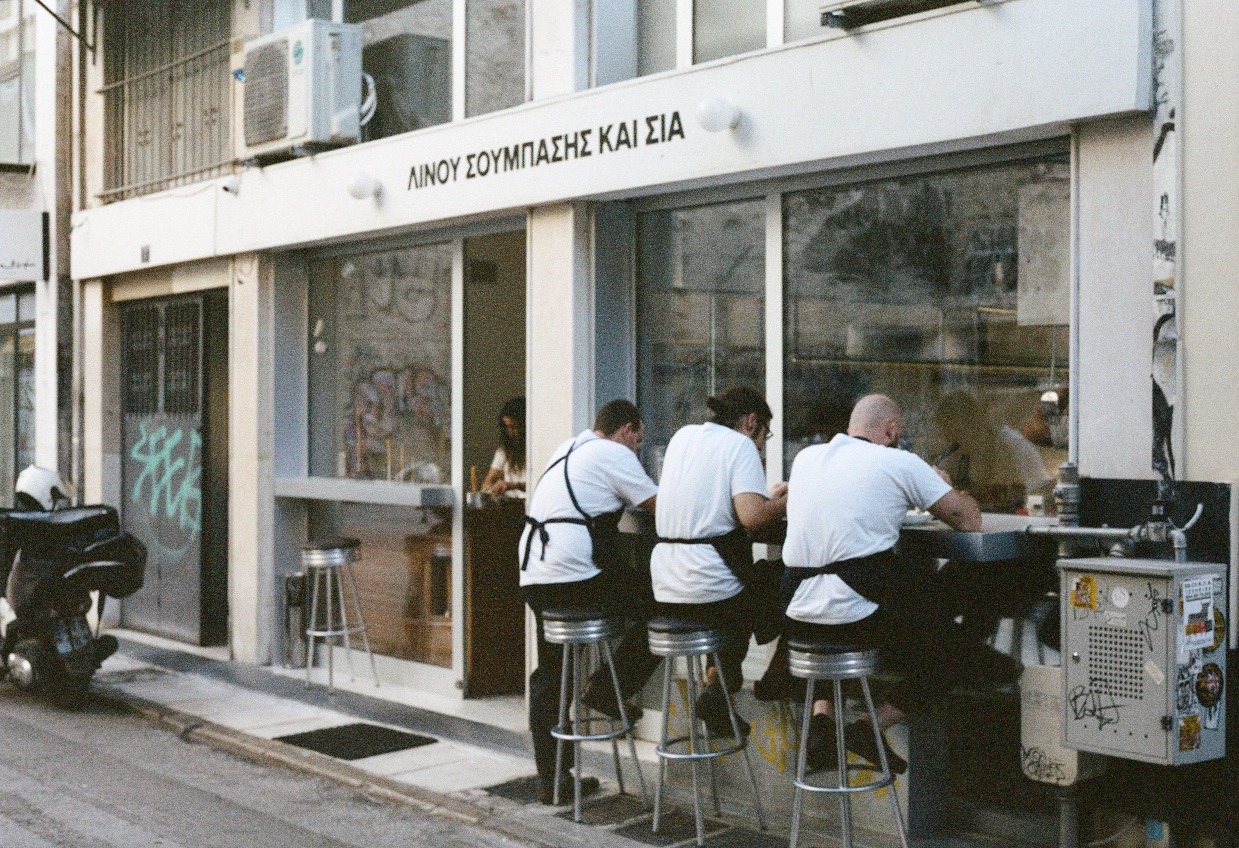 Three men sitting at a counter outside a restaurant with Greek writing, wearing black aprons, eating or drinking, with an indoor customer in the background, and a scooter parked on the street.