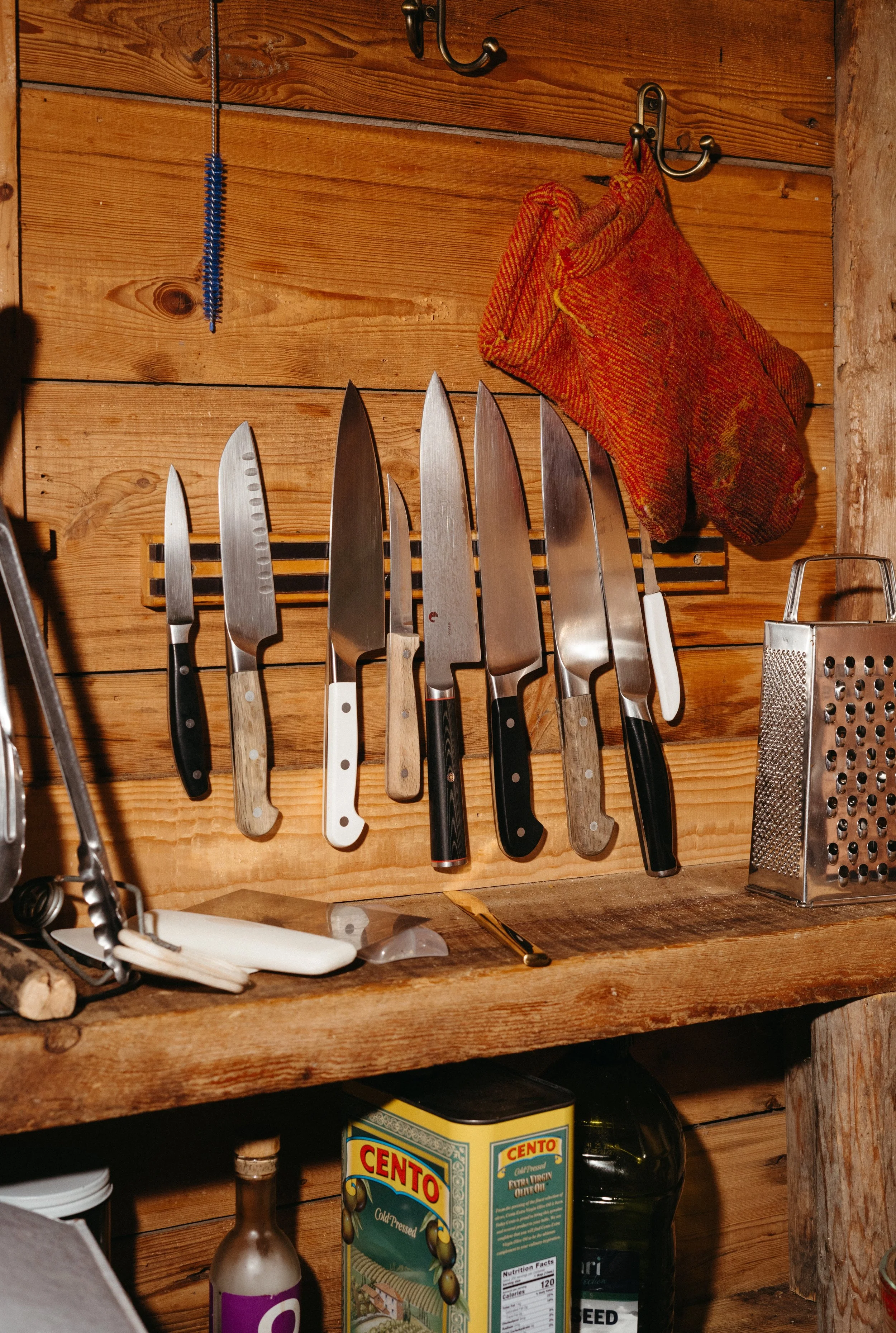 A wooden wall with a magnetic strip holding six kitchen knives with various handles, a blue brush hanging, and an orange oven mitt hanging on hooks. There is a grater to the right, some objects on the shelf below including a box of Cento tomato paste