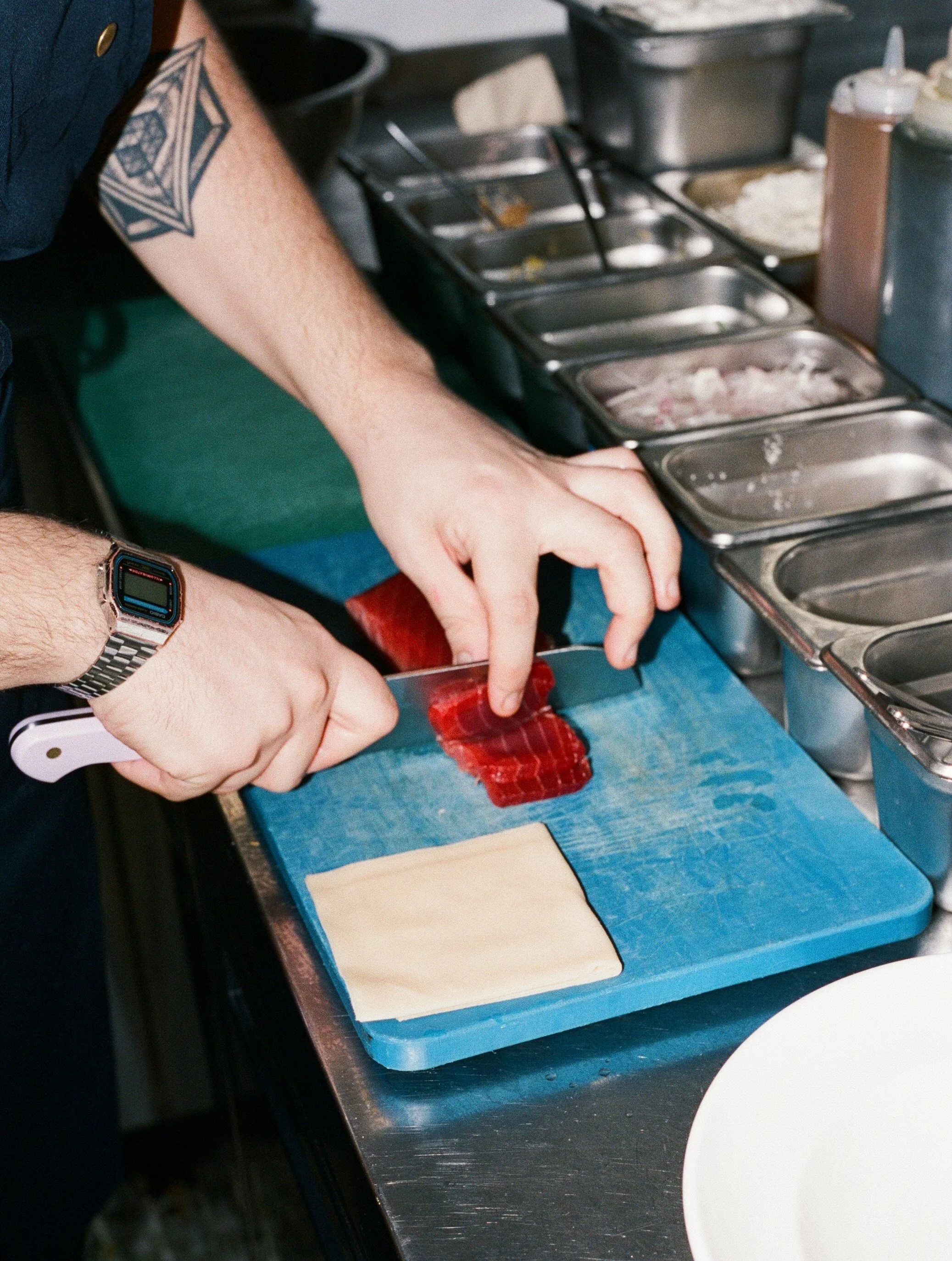 A person is slicing a piece of raw beef on a blue cutting board with a white-handled knife, with a piece of dough nearby. There are metal containers with various ingredients in the background.