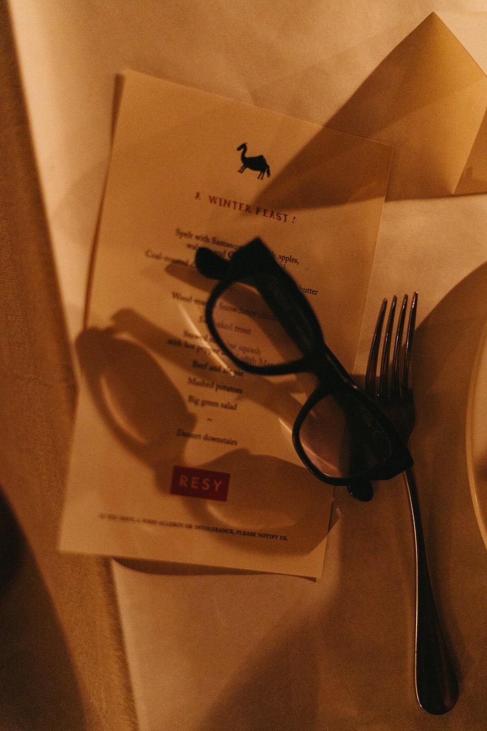 A folded card titled 'A WINTER FEAST!' with a silhouette of a goat at the top, a pair of black glasses, a fork, and a beige napkin on a table with warm lighting.