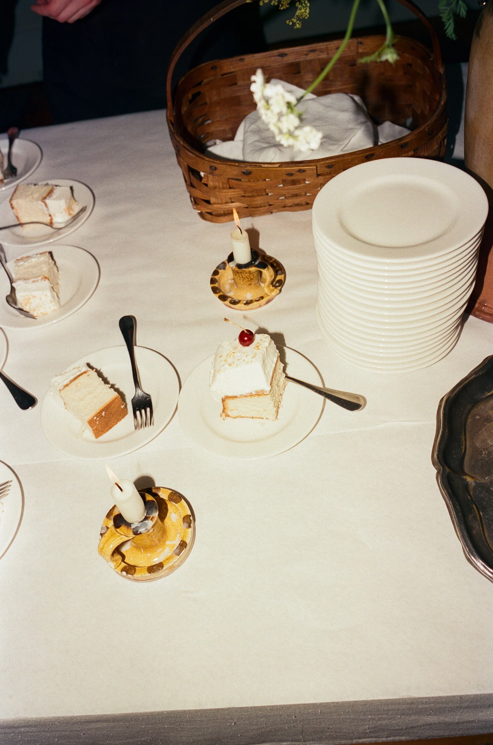 A table with multiple slices of cake on plates, some with forks, two lit candles in candle holders, a stack of white plates, a wicker basket, and a small pitcher with white flowers.