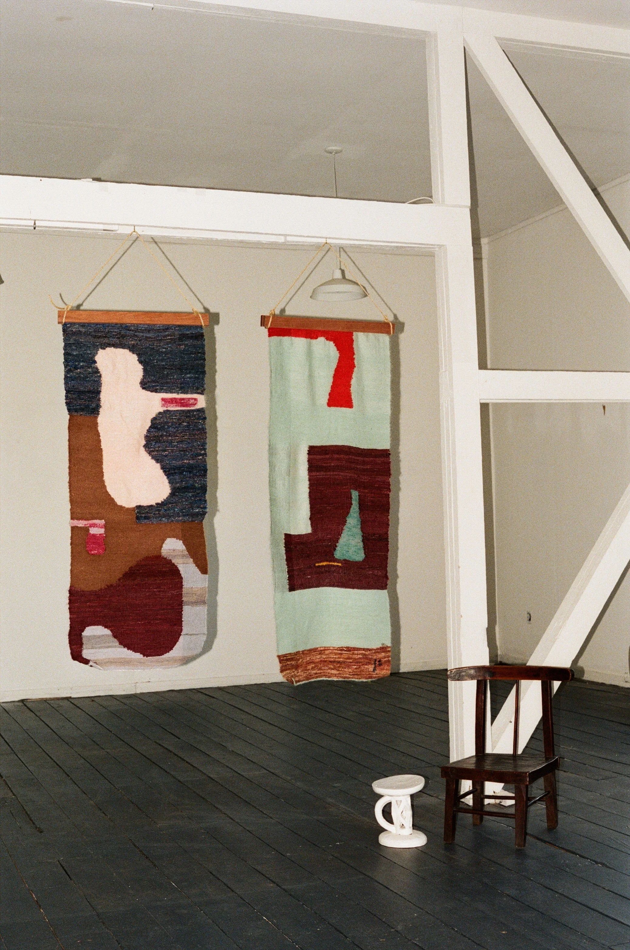 Two woven tapestry wall hangings with abstract designs hanging on a white wall in a room with dark wooden flooring, and a small wooden chair with a white ceramic stool nearby.