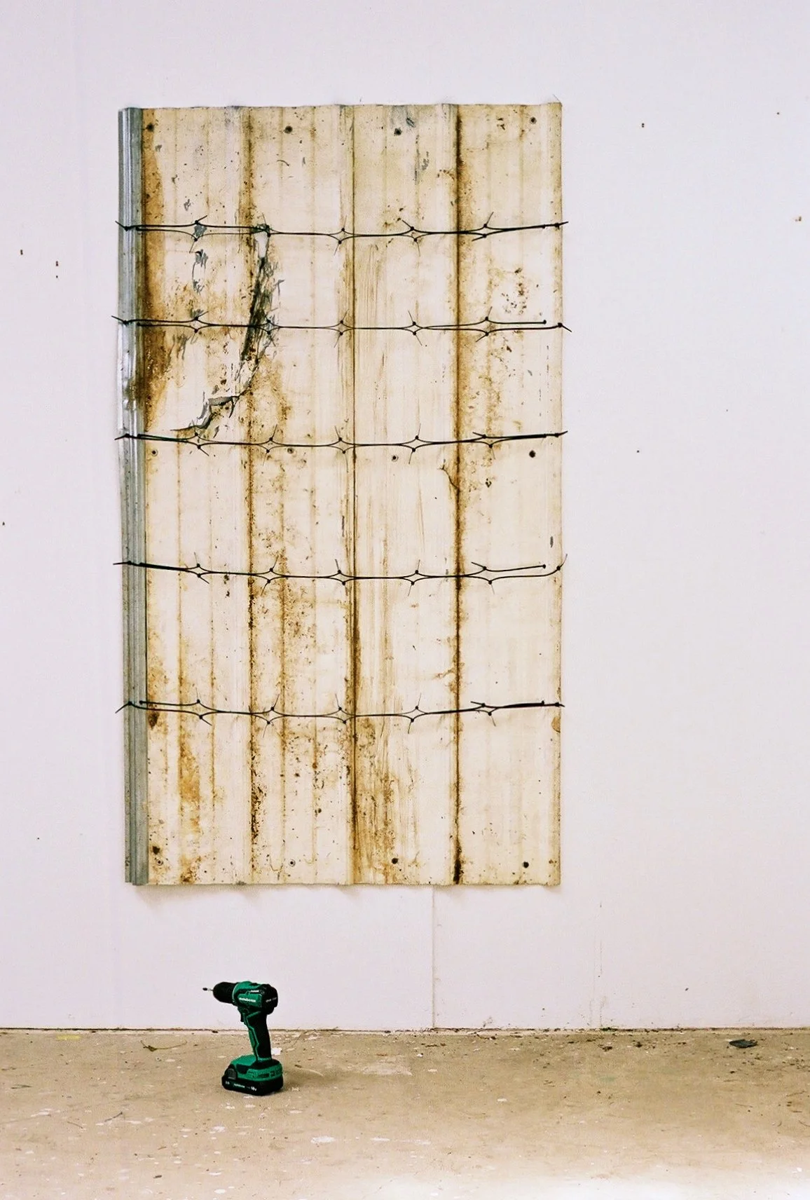 A wall with a large rusted, weathered metal panel and barbed wire attached to it. A power drill is on the floor in front of the wall.