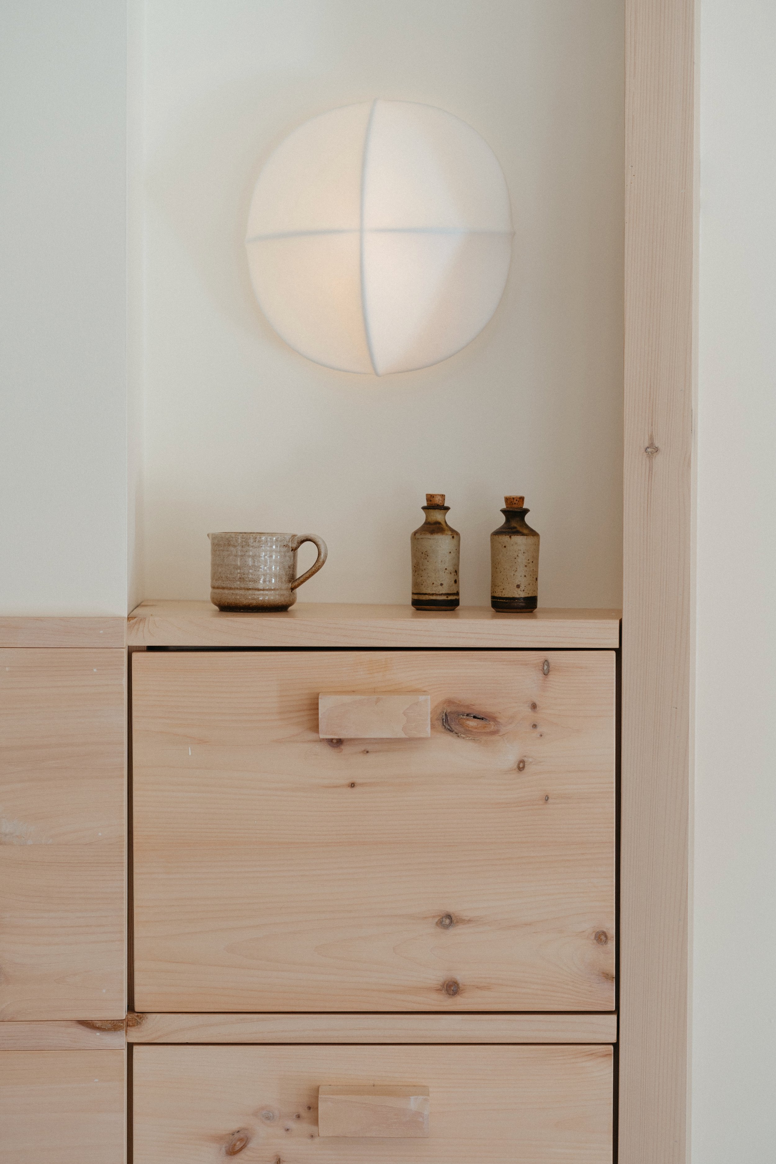 Minimalist wooden cabinet with a textured ceramic mug and two small glass bottles on top, illuminated wall light above.