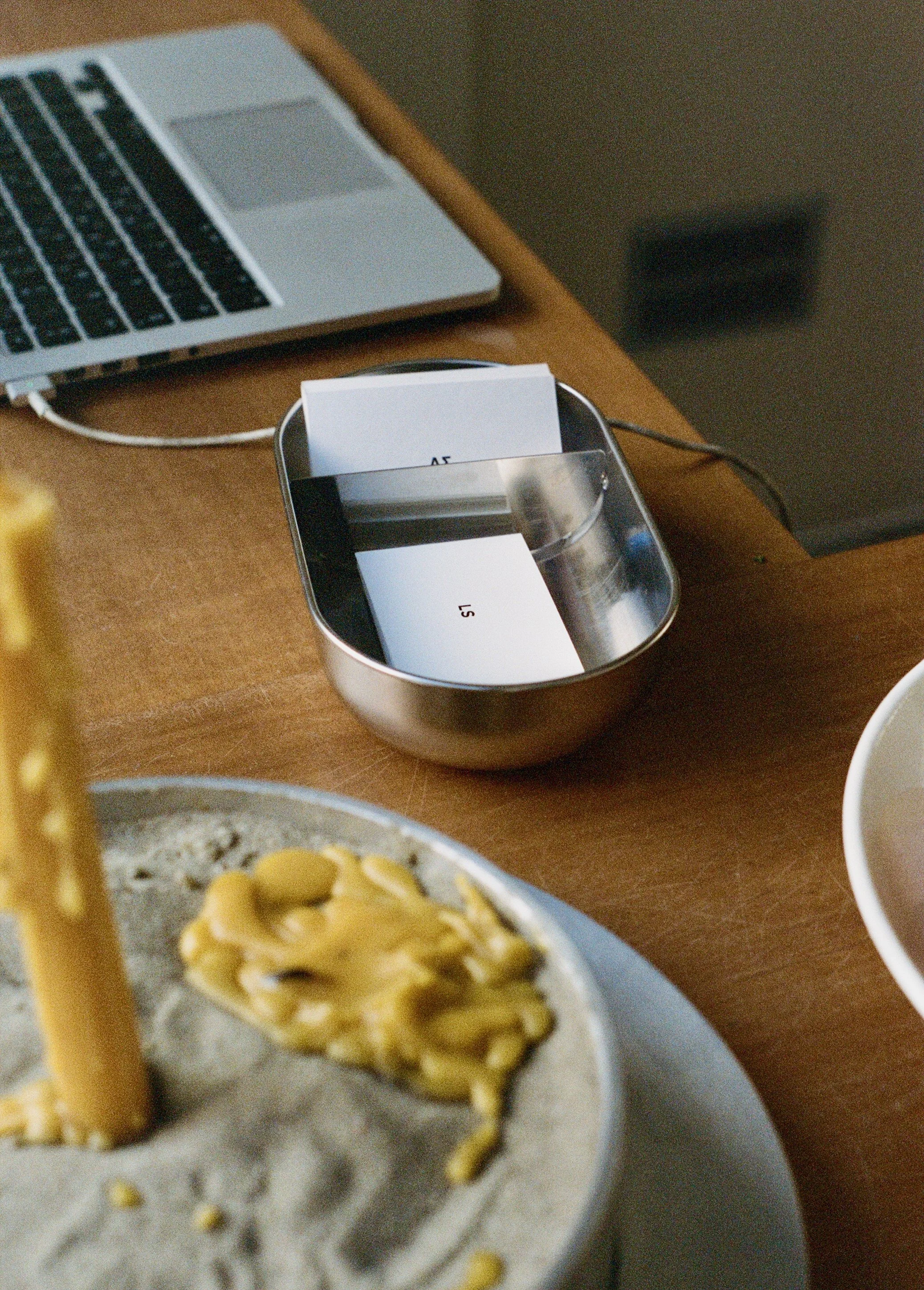A desktop scene with a laptop, a metallic container with tickets, a plate with pasta, and a white bowl on a wooden table.