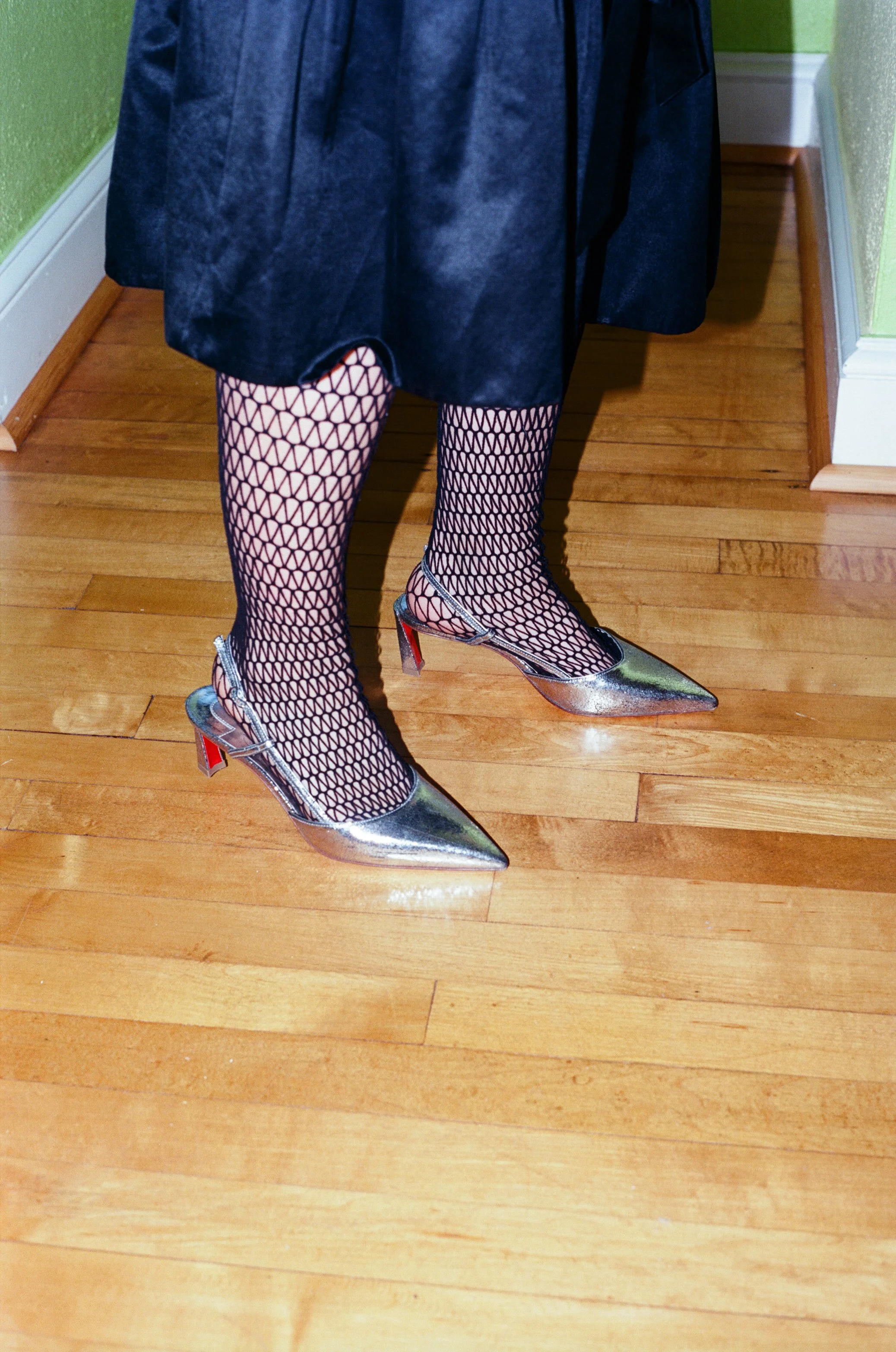 Person wearing fishnet stockings and metallic high-heeled shoes with a pointed toe, standing on a hardwood floor next to a green wall.