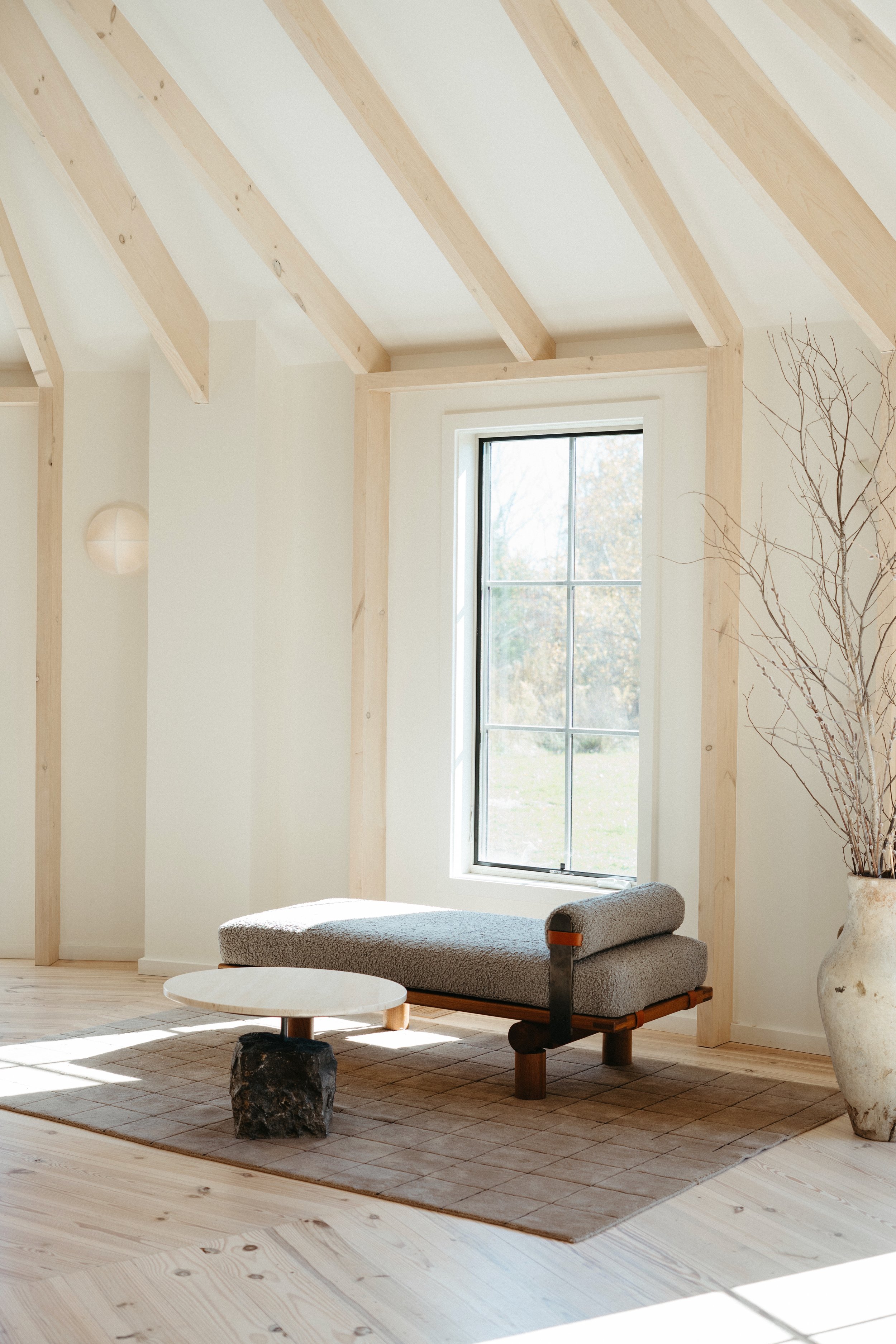 Minimalist living room with natural wood beams, a large window, and a modern lounge chair on a neutral rug.
