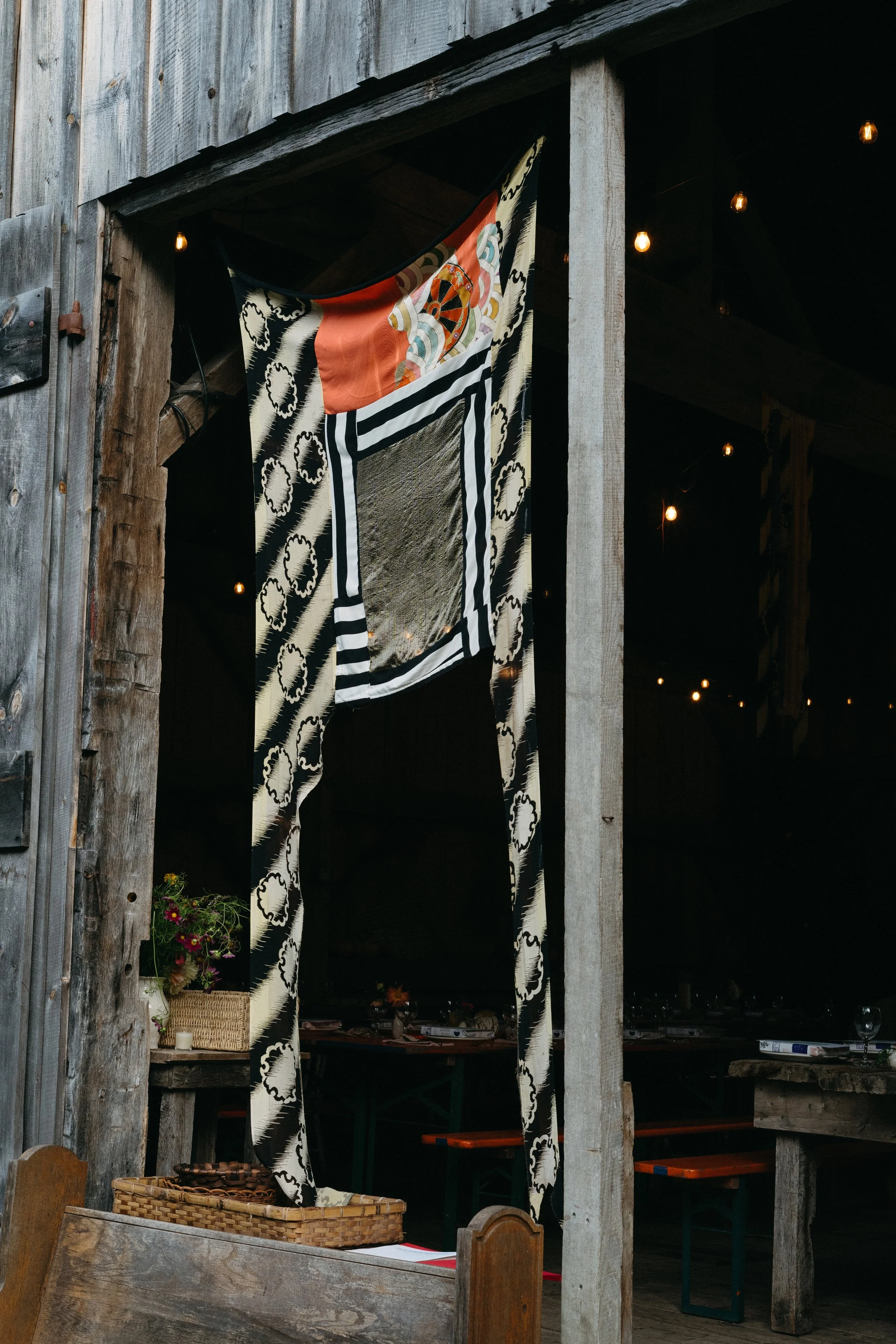 Inside a rustic barn or restaurant with a wooden structure, decorated with a patterned black and white curtain and a quilt that features a design of a ship's wheel and sailboat, illuminated by small hanging string lights.