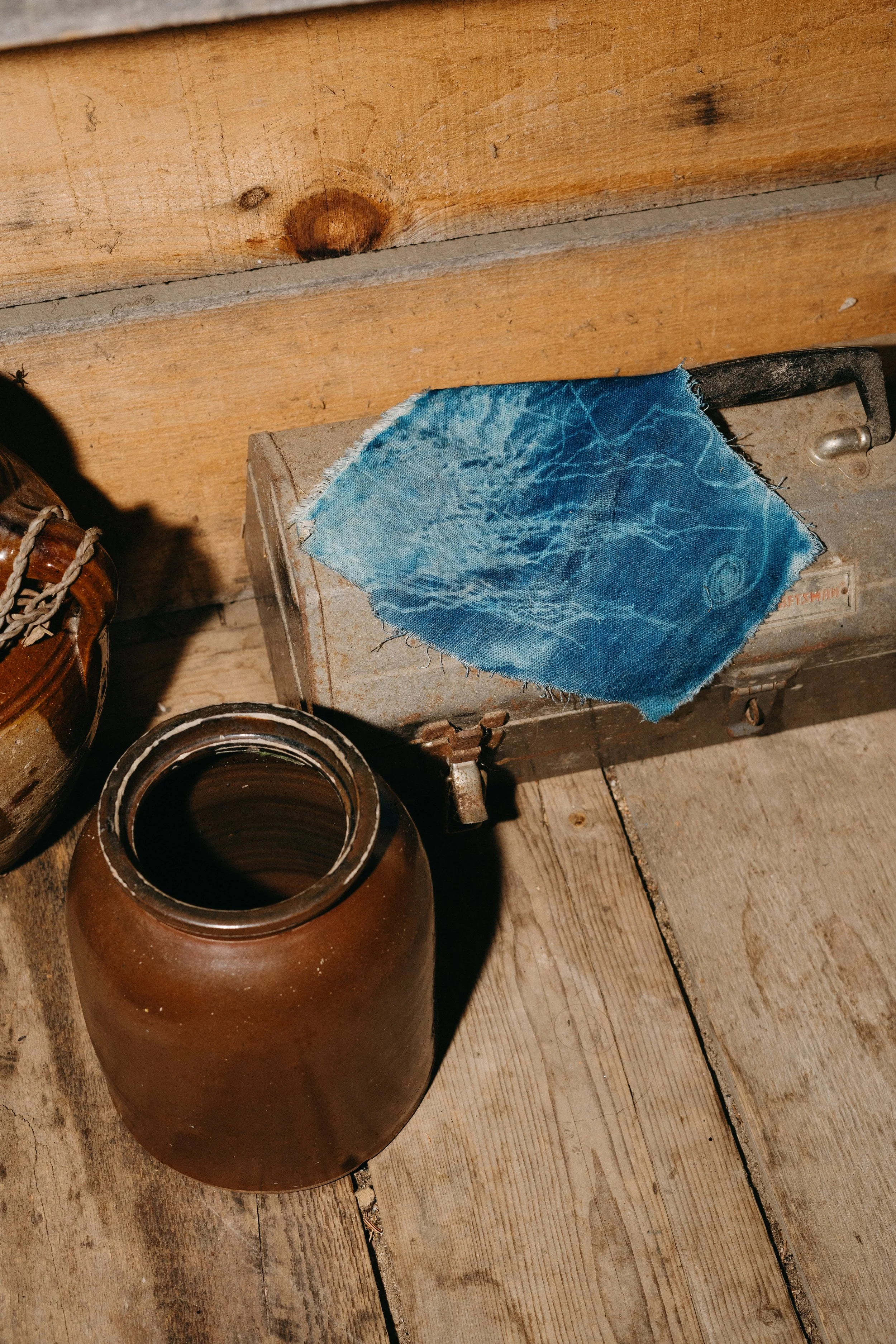 An old wooden floor with a brown ceramic jar and an old metal box with a piece of blue fabric on top.