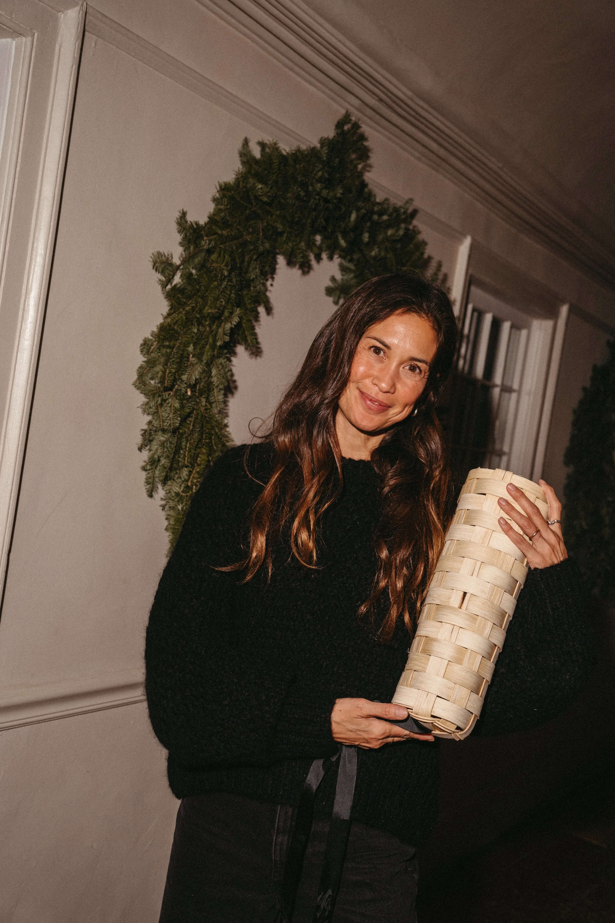 Woman with long brown hair in black sweater holding a woven basket on a decorated porch with greenery