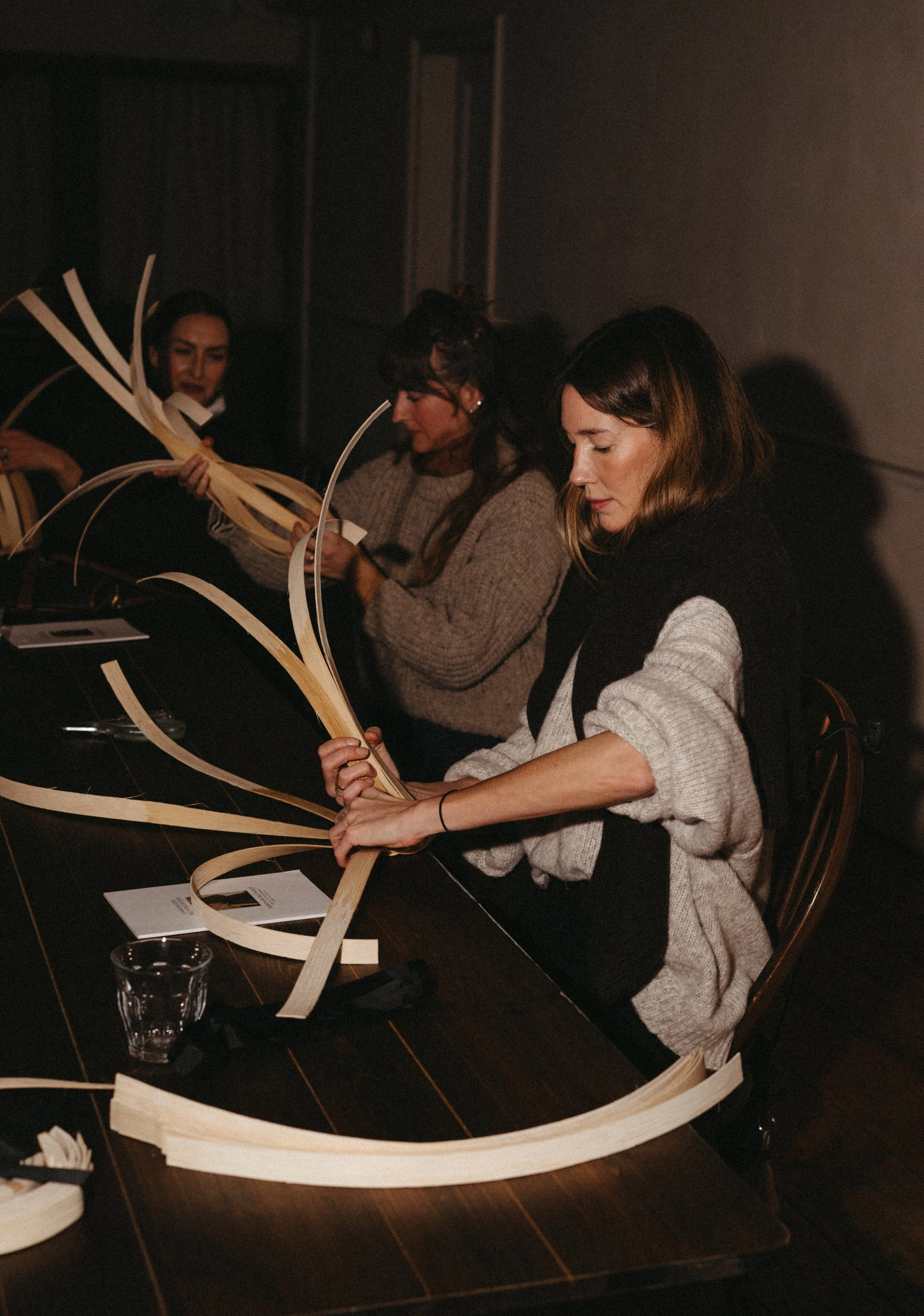 Three women sit at a table, working with long strips of wood or paper, in a dimly lit room.