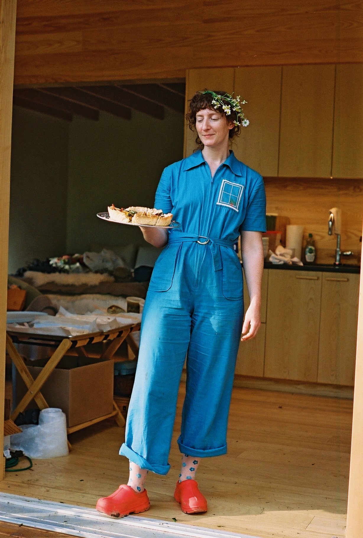A woman with curly brown hair, wearing a blue jumpsuit, floral headband, red clogs, and patterned socks, standing indoors with a plate of food in her hand, in a cozy wooden kitchen or living area.