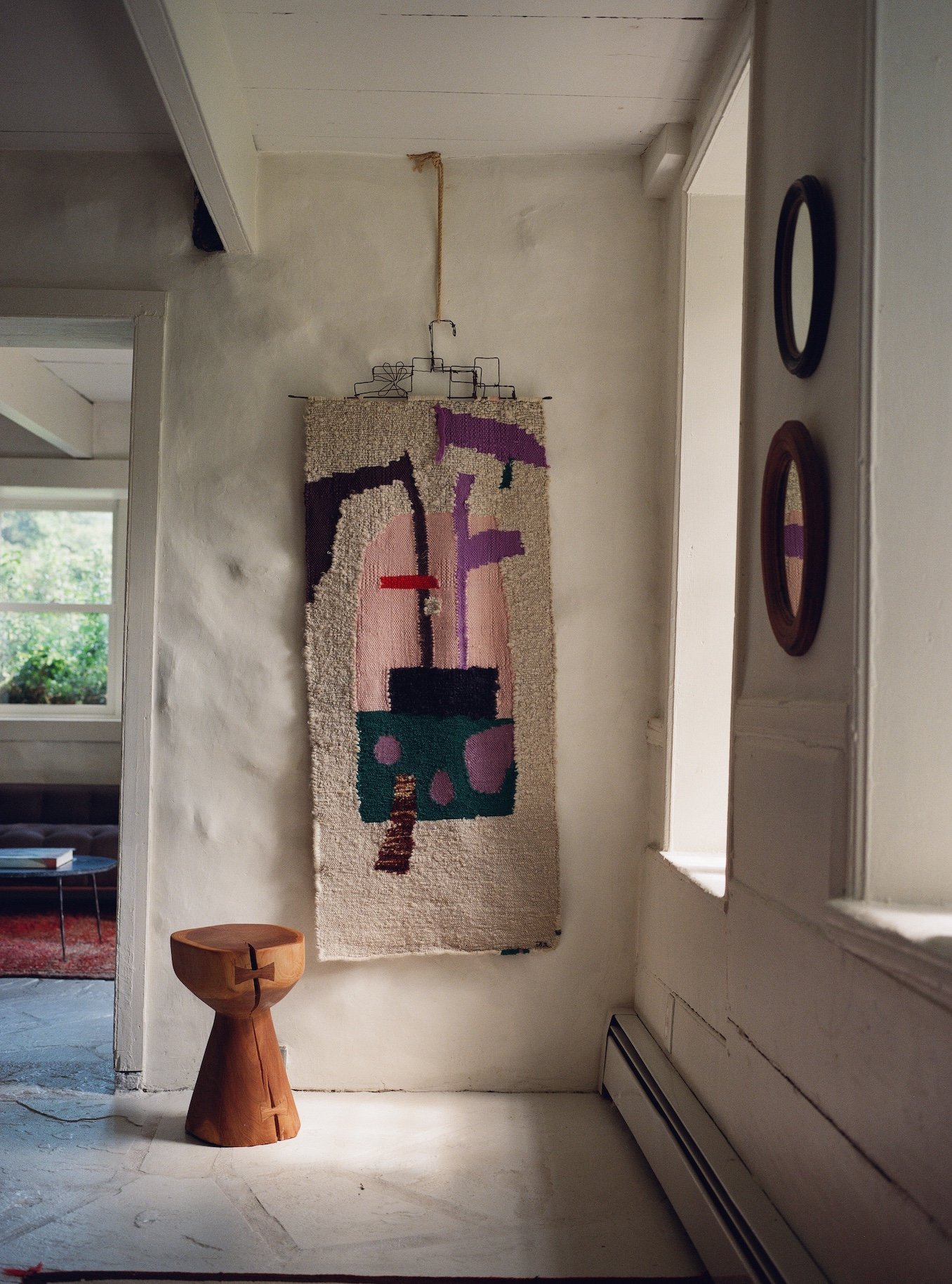 A woven wall hanging artwork in abstract design, with dark pink, black, purple, and green accents, hangs on a white wall. A small wooden stool with a rounded top and carved base stands in front of the wall, next to a window with white trim in a cozy 