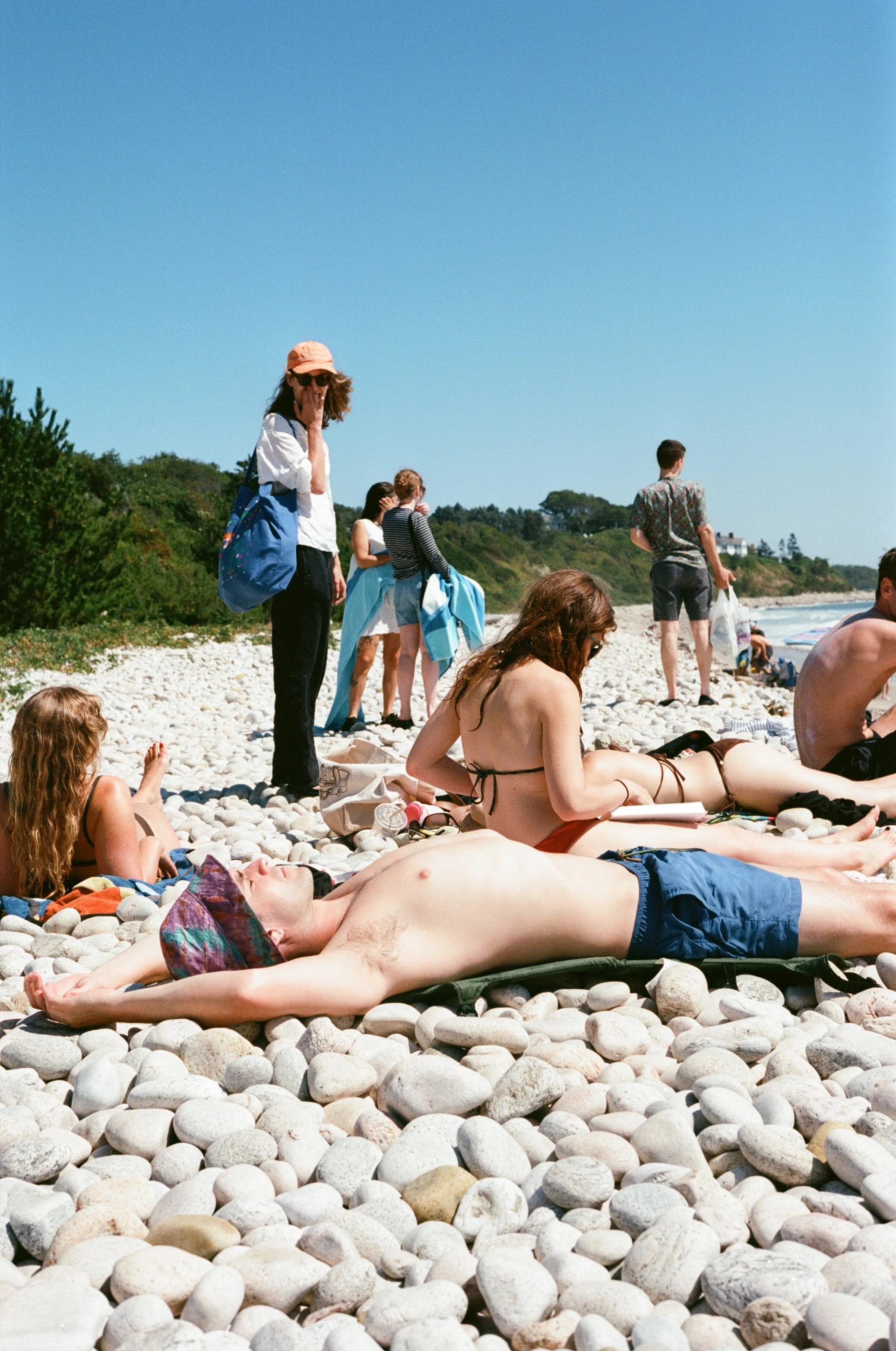 People relaxing and sunbathing on a pebble beach on a sunny day, with some standing and others lying down on the rocks.