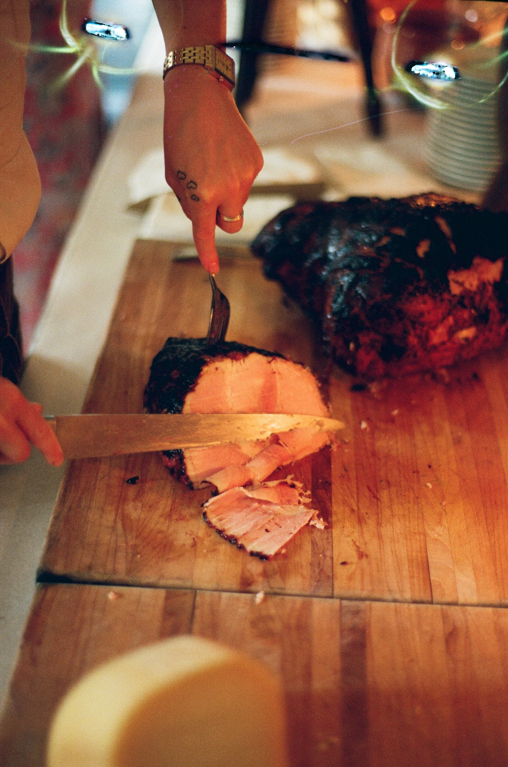 Person slicing cooked meat on a wooden cutting board with a knife, with other cooked meat in the background.