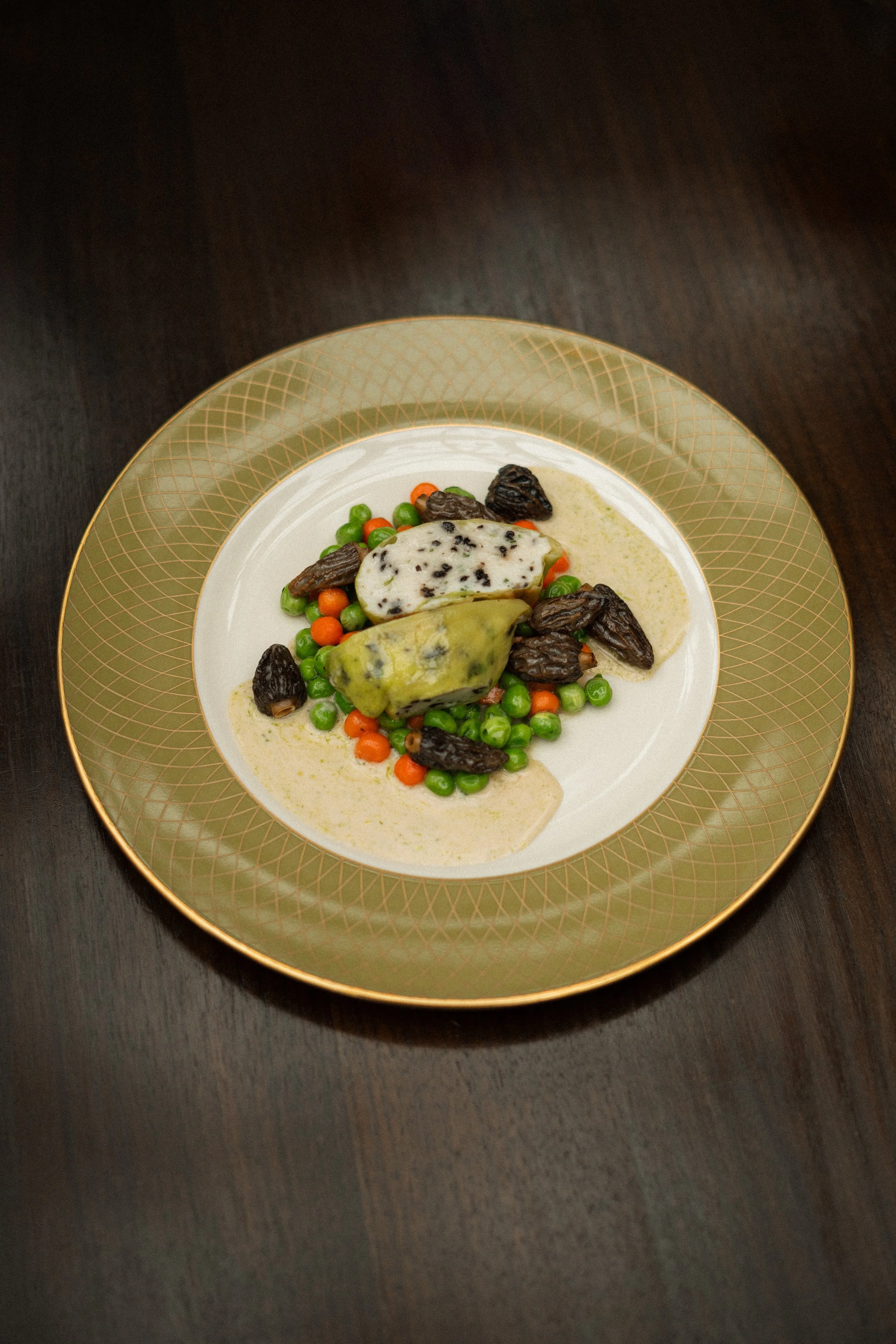 A plated dish with three types of cheese, green peas, orange carrots, glazed dates, served with a light sauce on a decorative green and gold-rimmed plate.