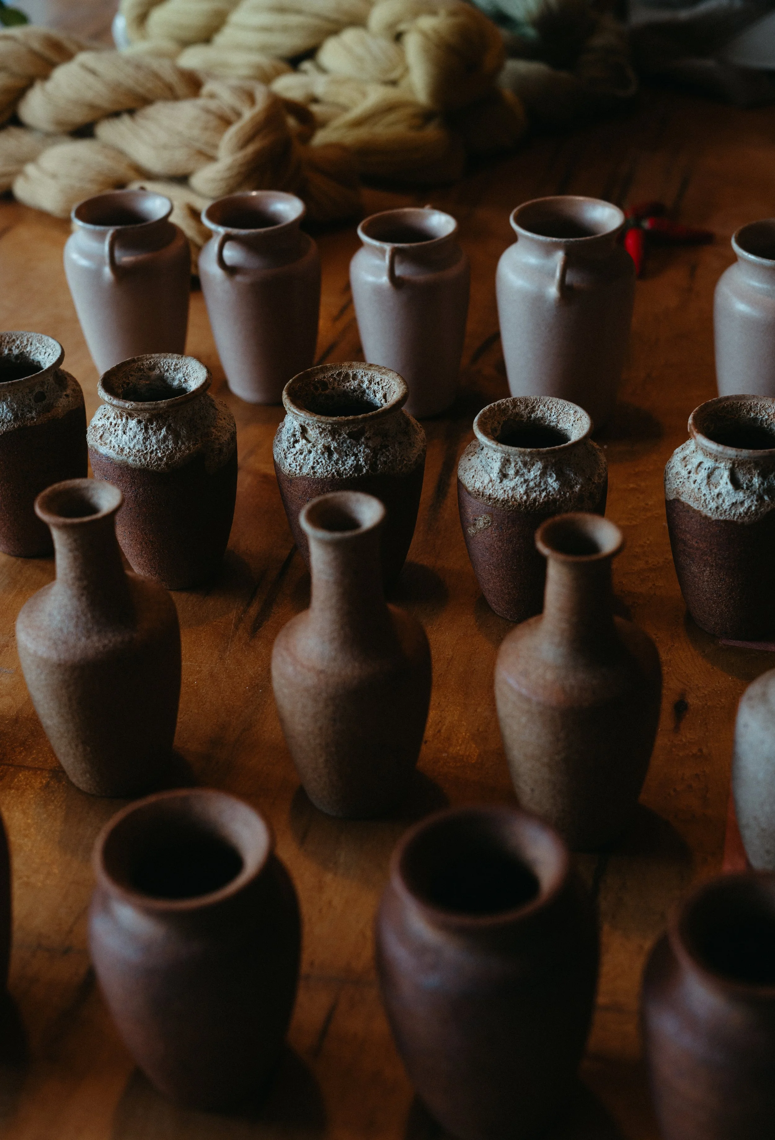 Multiple small pottery vases and jugs on a wooden surface, some filled with dark liquid, with braided rope and fabric in the background.