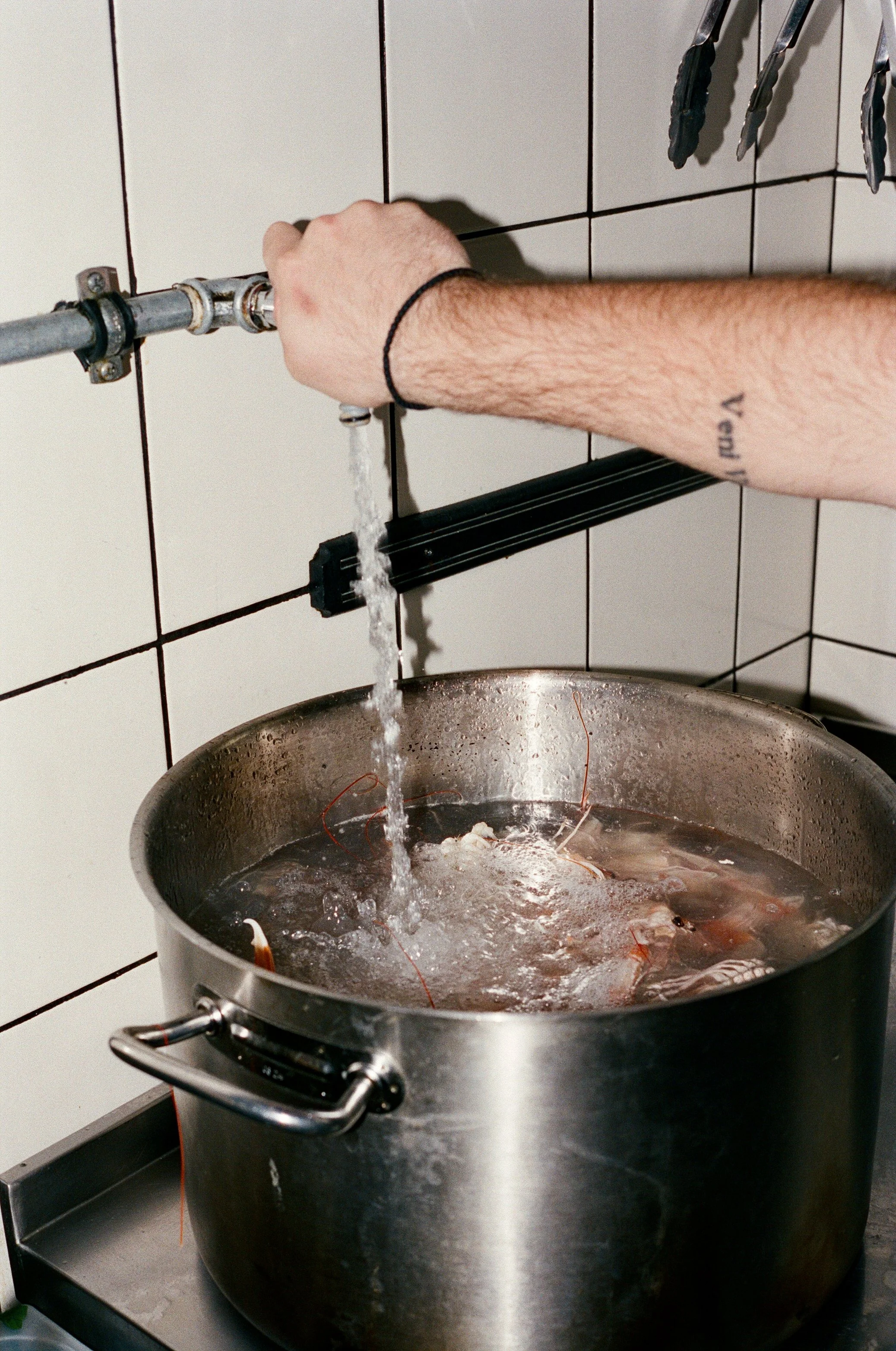 A person holding a pipe attached to a water source over a large stainless steel pot with boiling water and shrimp, in a tiled kitchen.