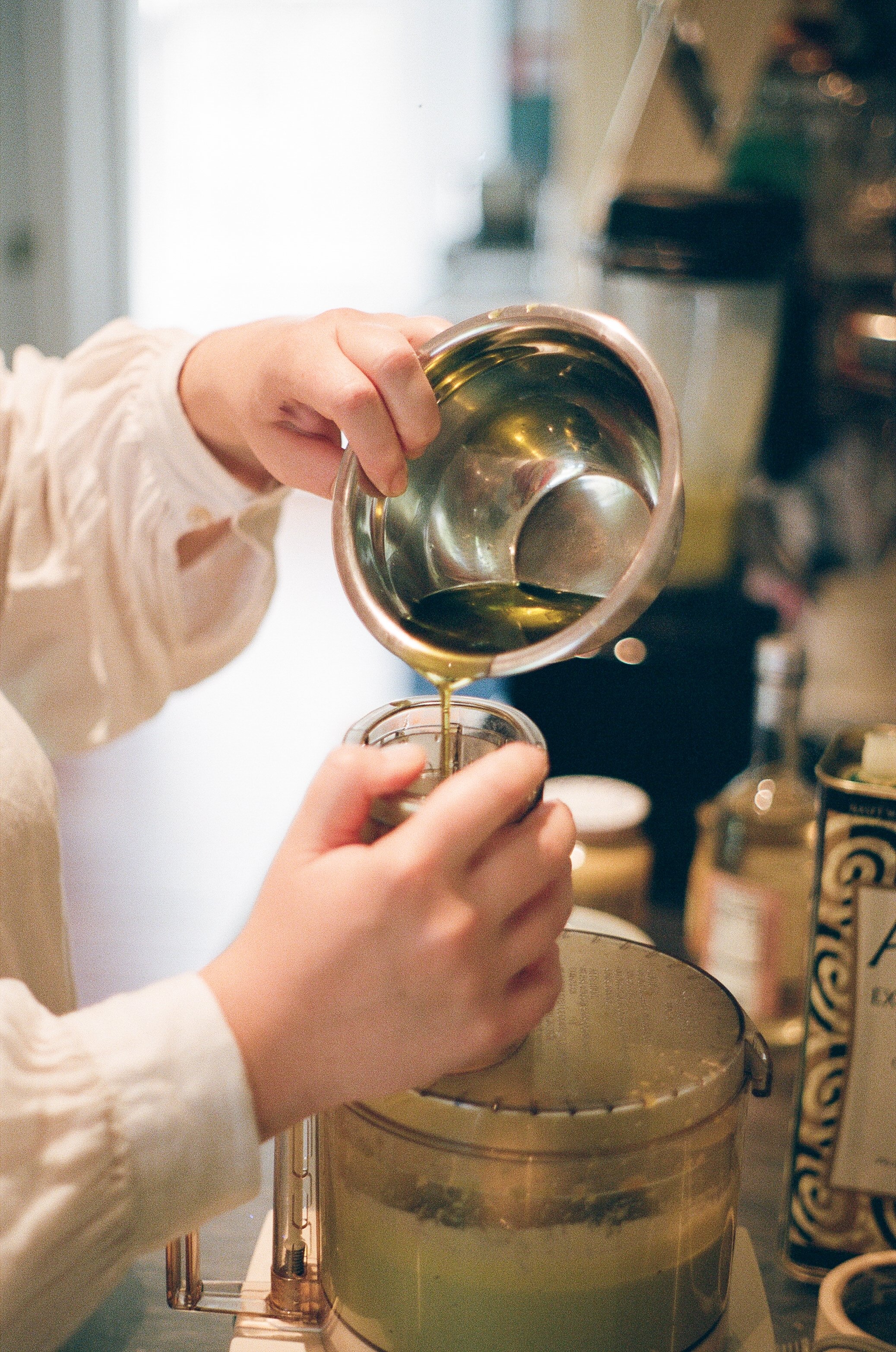 Person pouring olive oil from a stainless steel container into a small glass jar in a kitchen.