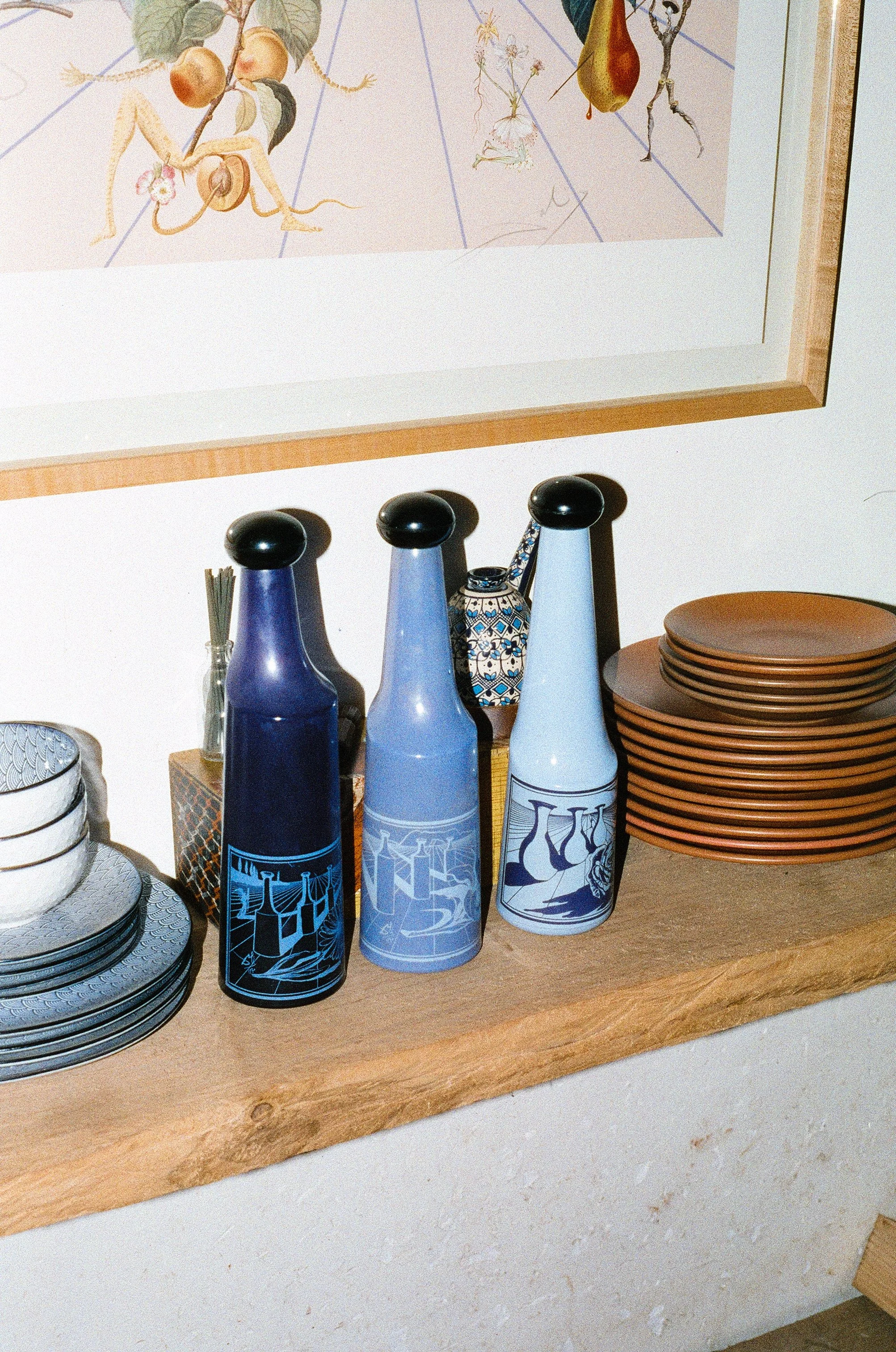 Three decorative bottles with black caps and blue shades, a small teapot with a blue and white pattern, a stack of brown plates, and a few small bowls with blue and white patterns on a wooden shelf.