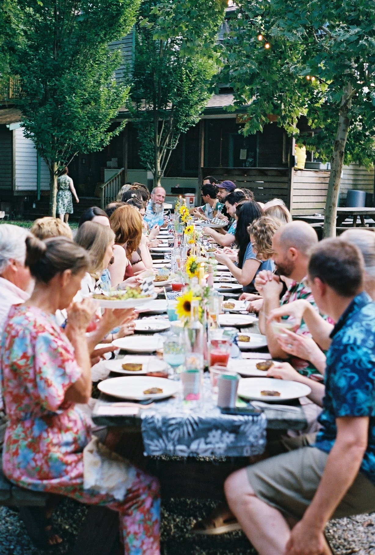 People gathered around a long outdoor dining table with sunflower centerpieces at a backyard party.