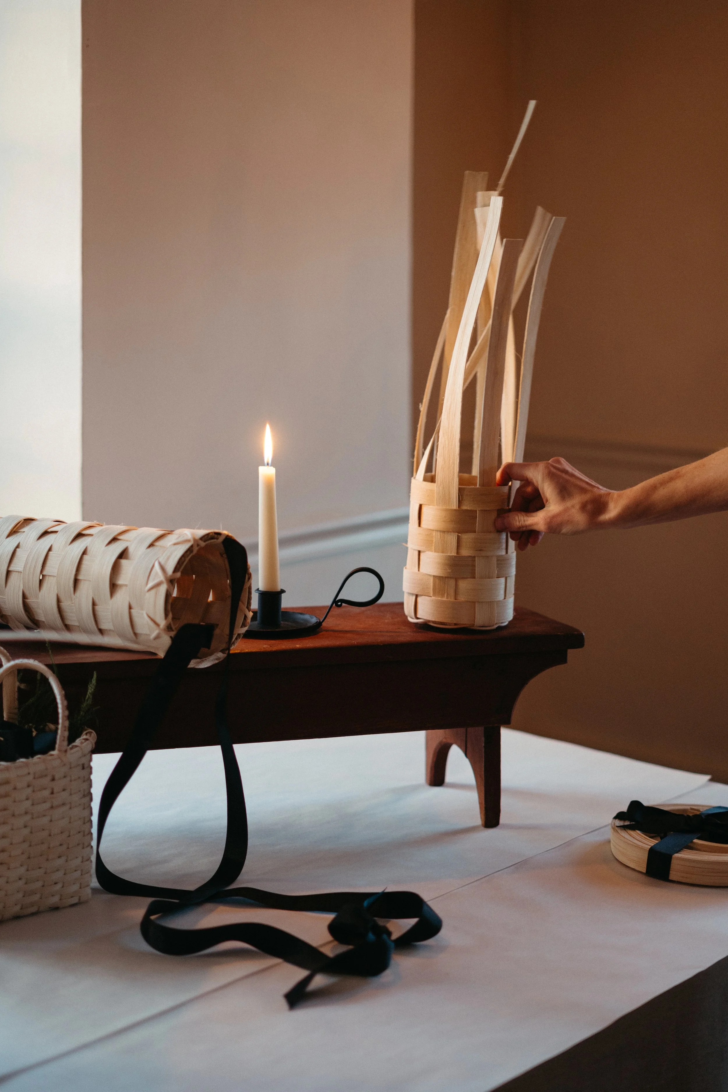 A person arranging a woven basket on a wooden table, with a lit candle and a rolled-up woven item nearby, in a cozy indoor setting.