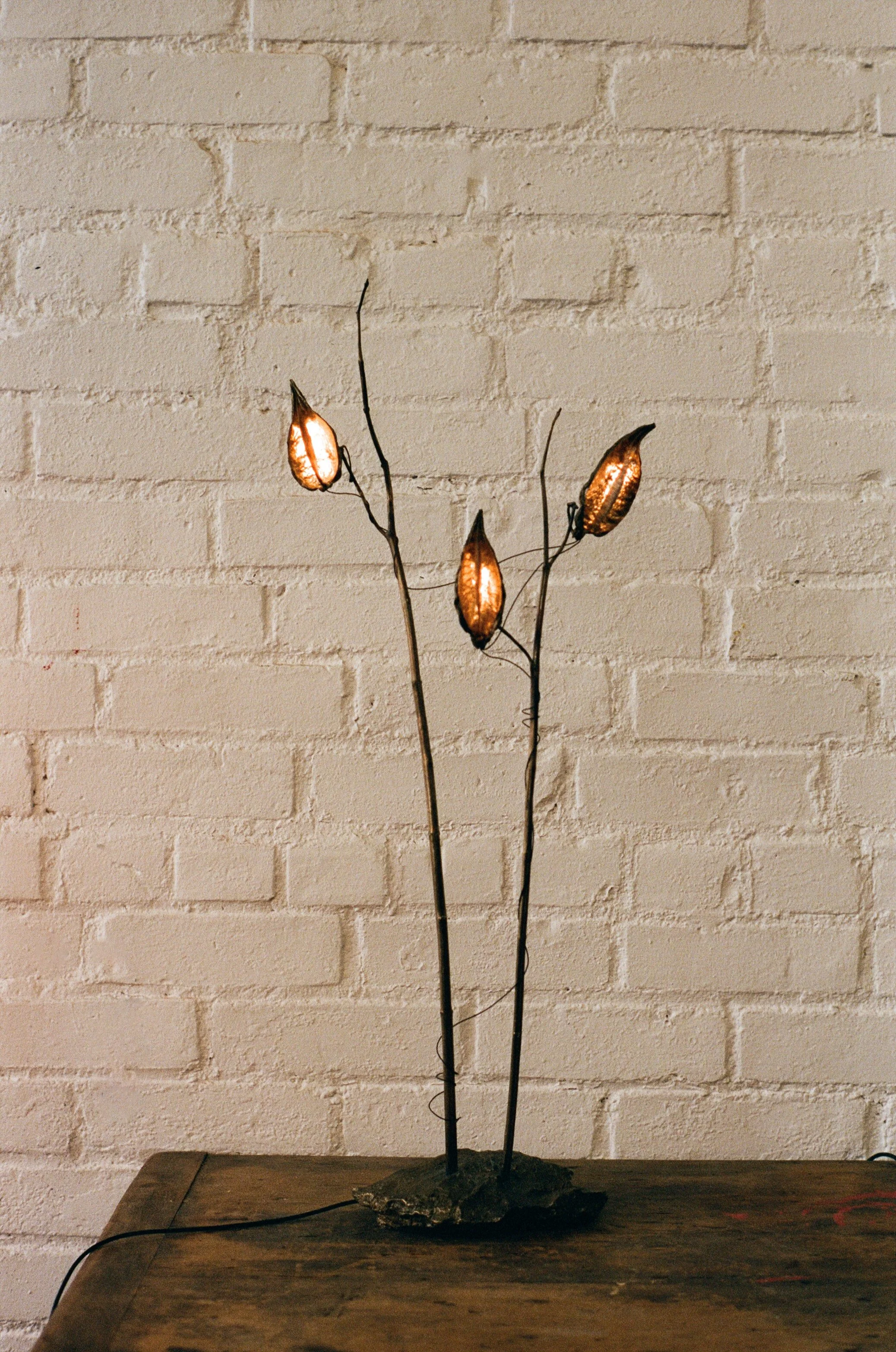 Decorative table lamp resembling a floral sculpture with five leaf-shaped light fixtures on thin, curved branches, placed on a wooden table against a brick wall.