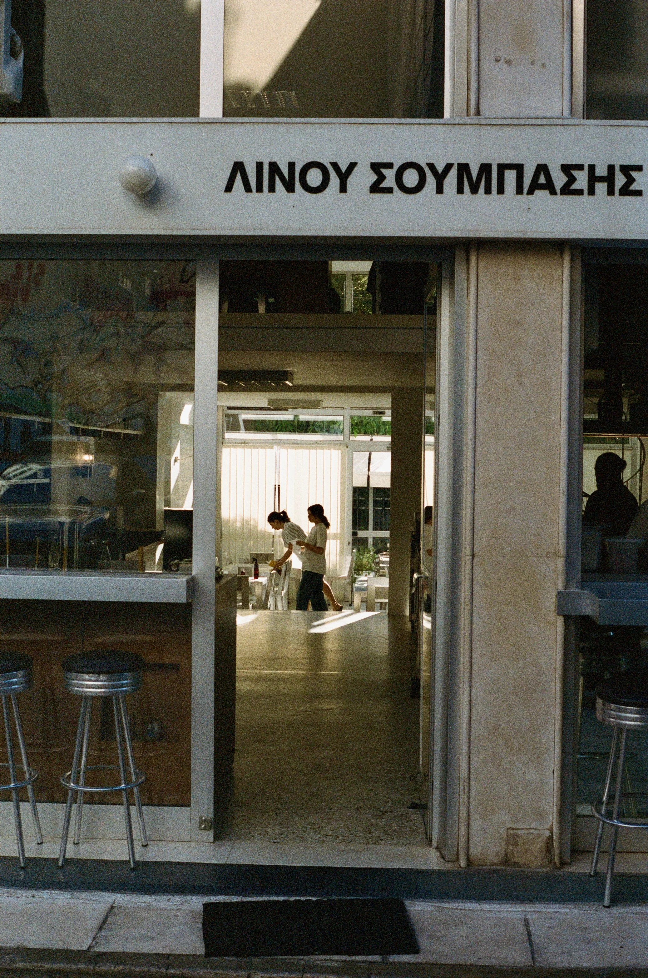 View of a restaurant entrance with Greek signage, showing two women inside preparing food or drinks, with barstools outside and natural light illuminating the interior.