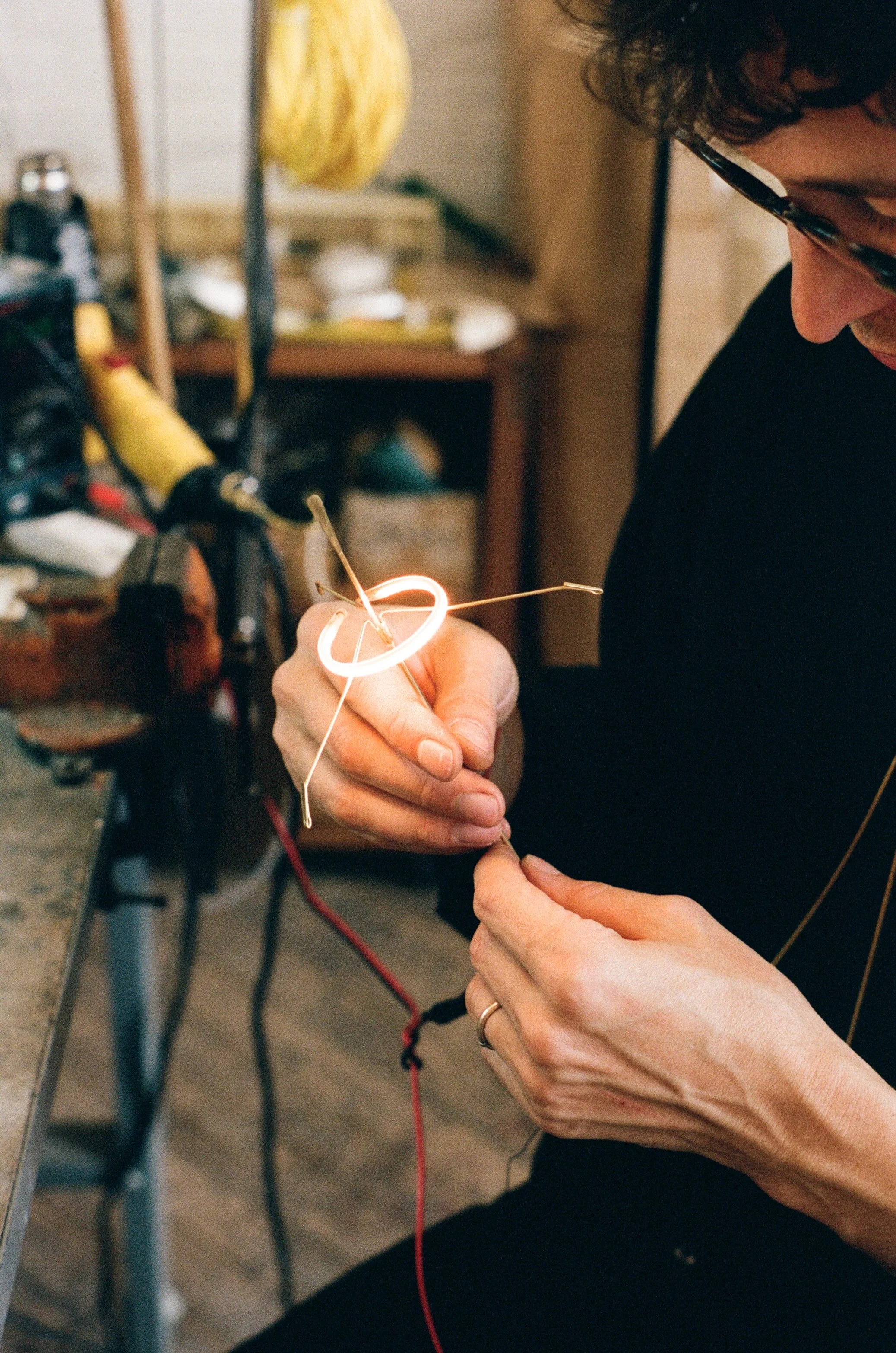 A person working with a small flame near a workshop table, holding a glowing ring with metal wires attached, surrounded by tools and equipment.