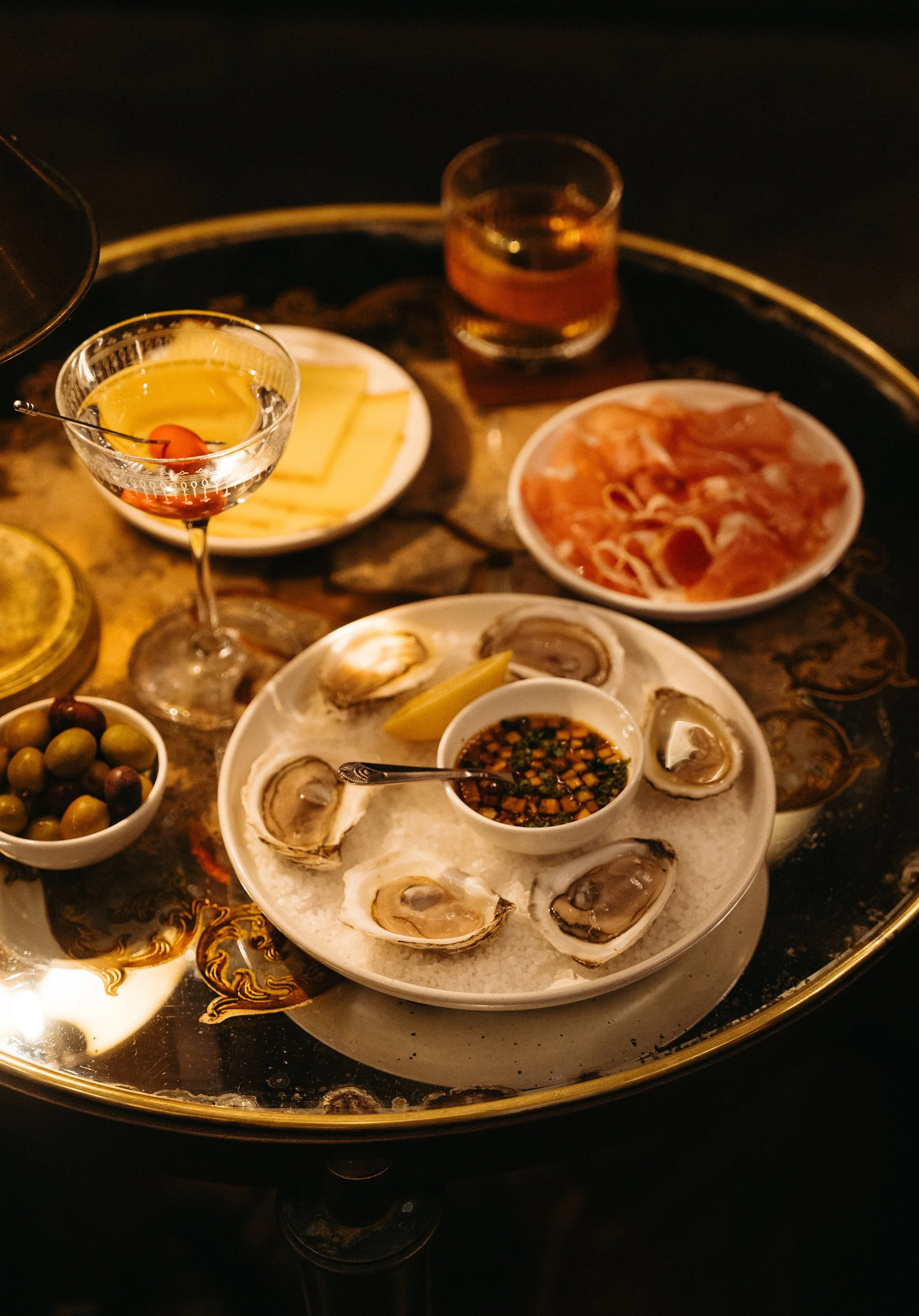 A tray with a seafood appetizer of oysters, green and purple olives, sliced cheese, and small dish of dipping sauce. Additional dishes include a plate of sliced ham, a bowl of cheese, and a glass of wine. The scene appears to be in a dimly-lit restau