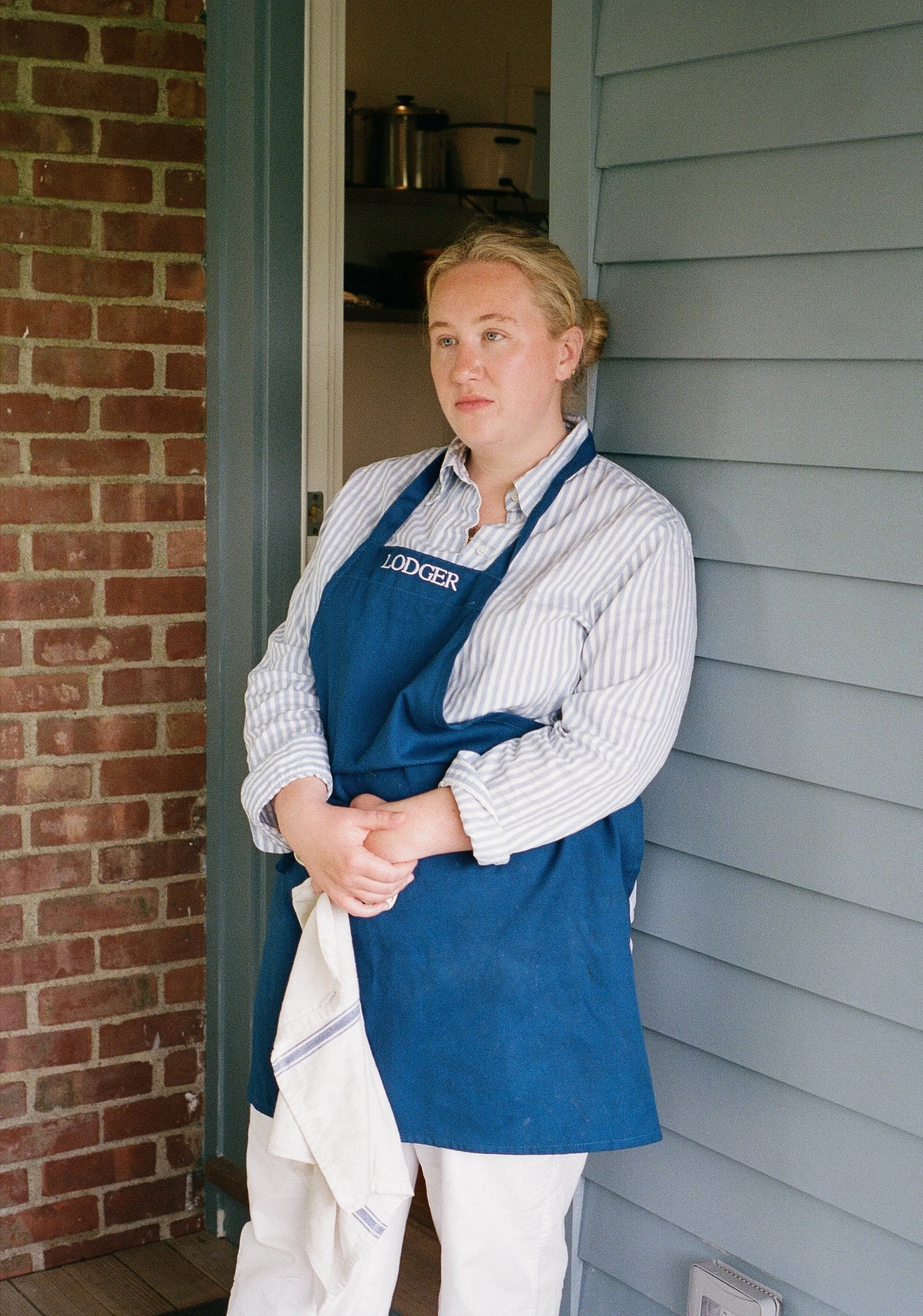 A woman wearing a blue apron with the name "Lodger" embroidered on it, standing outside a house with blue siding and a brick wall, looking to the side with a serious expression.
