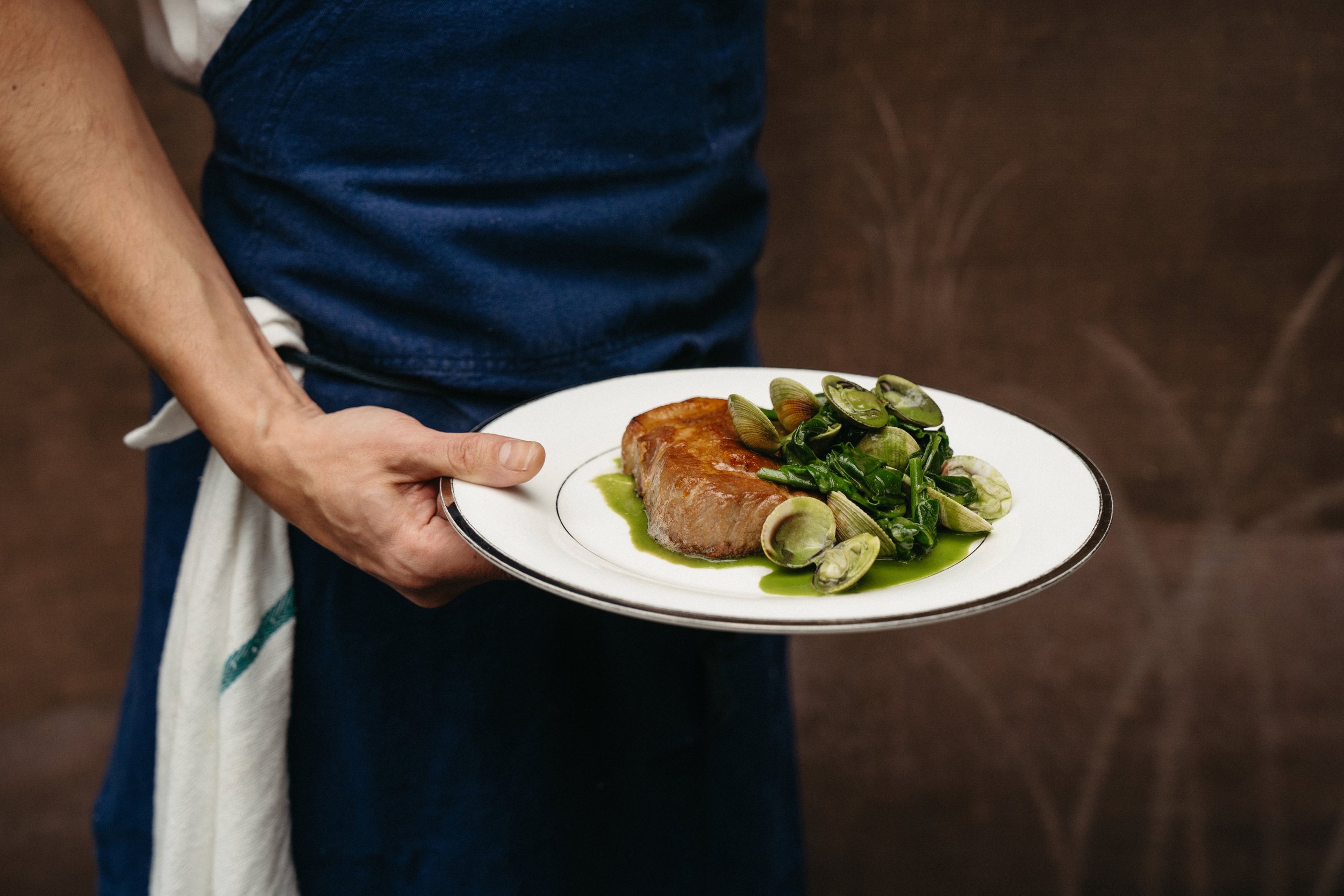 Person in a blue apron holding a white plate with a cooked piece of meat topped with green vegetables and surrounded by a green sauce.