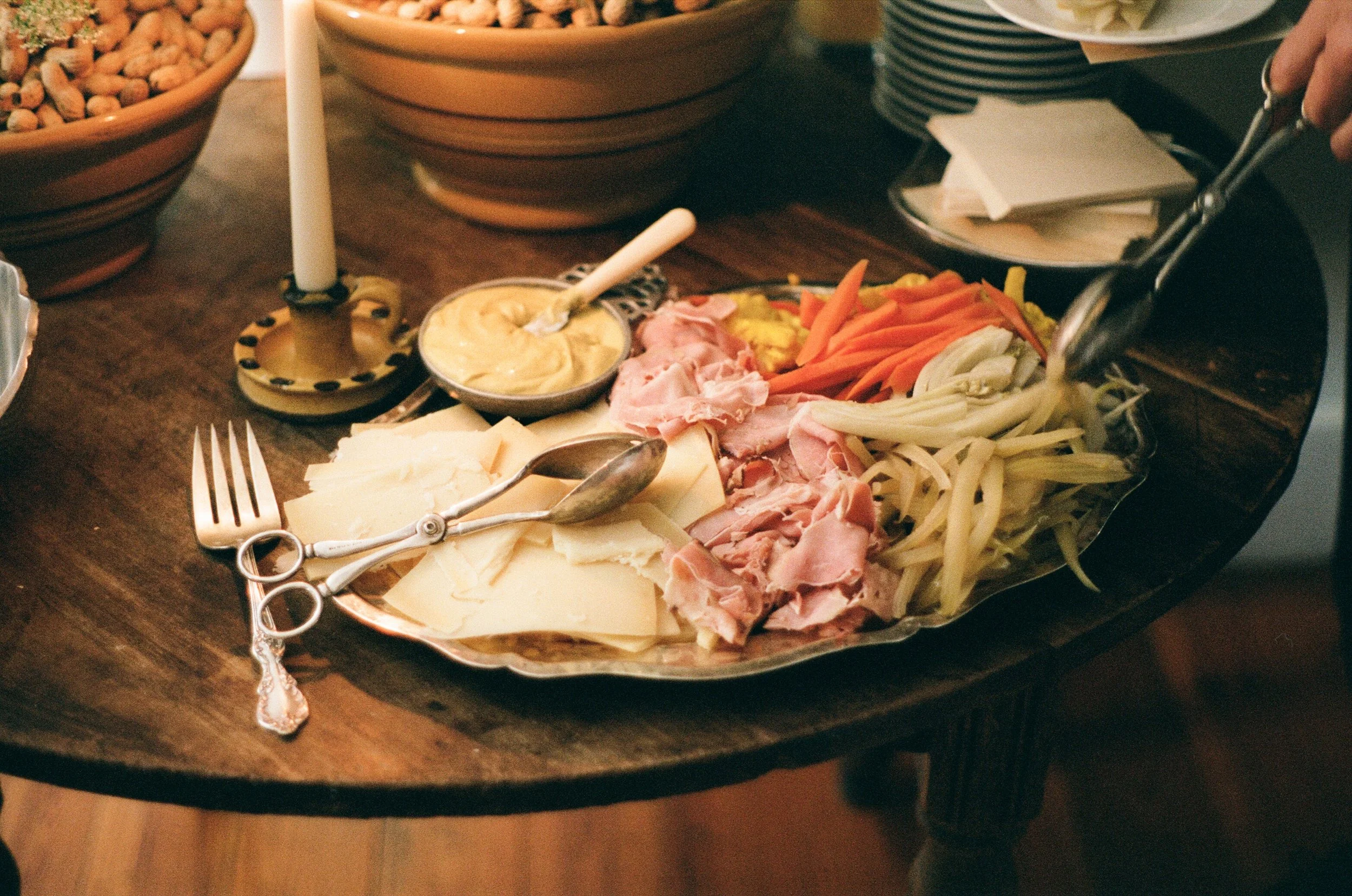 A cheese and meat platter with sliced cheeses, ham, turkey, and shredded vegetables, accompanied by a bowl of mustard or sauce, on a wooden table.