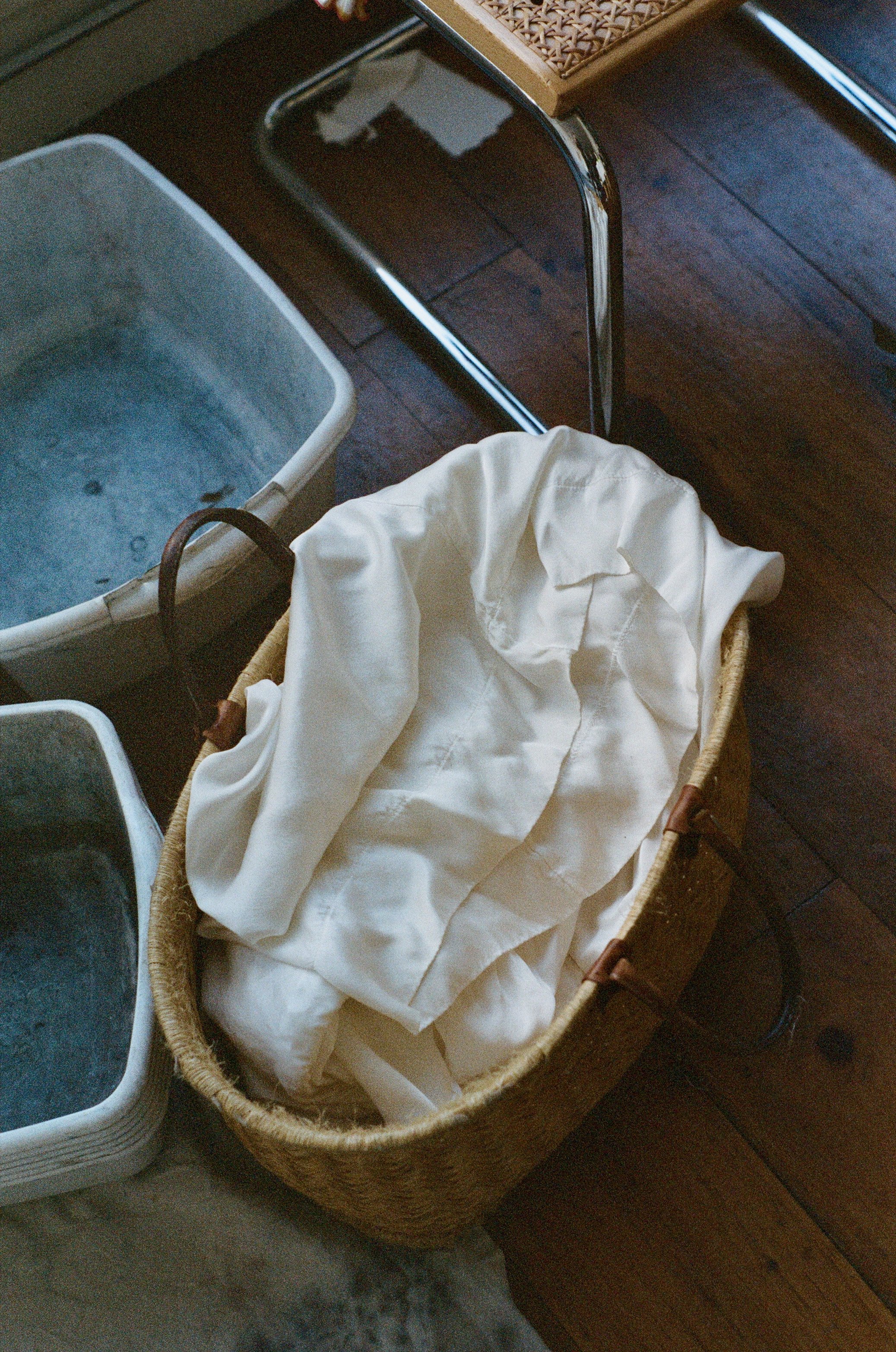 A woven basket containing white fabric or clothing, placed on a wooden floor next to metal tubs filled with water and a wooden table, indicating a laundry or washing area.