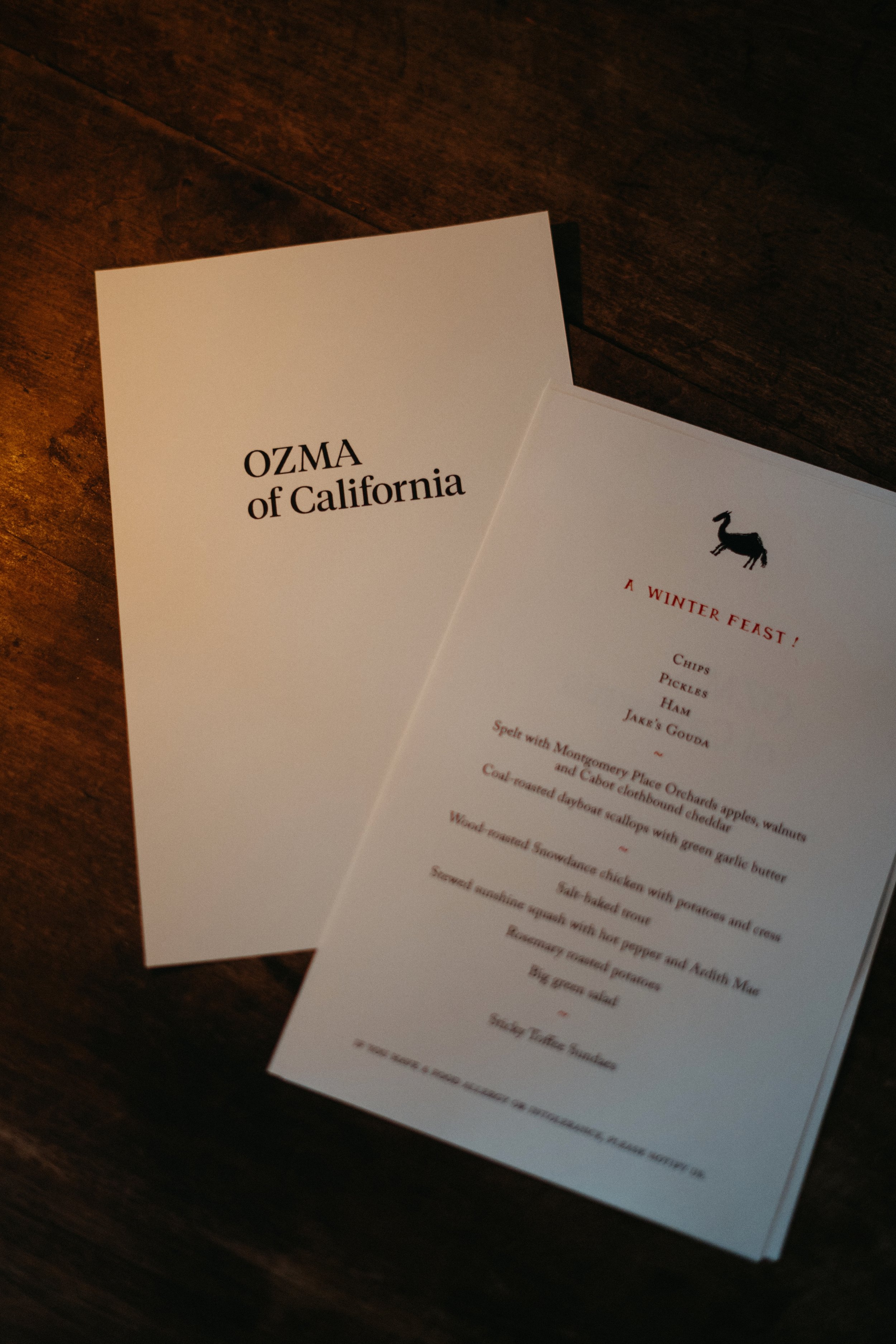 A menu titled 'A Winter Feast' with a black silhouette of a goat or similar animal at the top, featuring dishes like chips, pickles, ham, Jake's Gouda, and other traditional holiday foods, placed on a wooden table. The menu also includes a pamphlet o