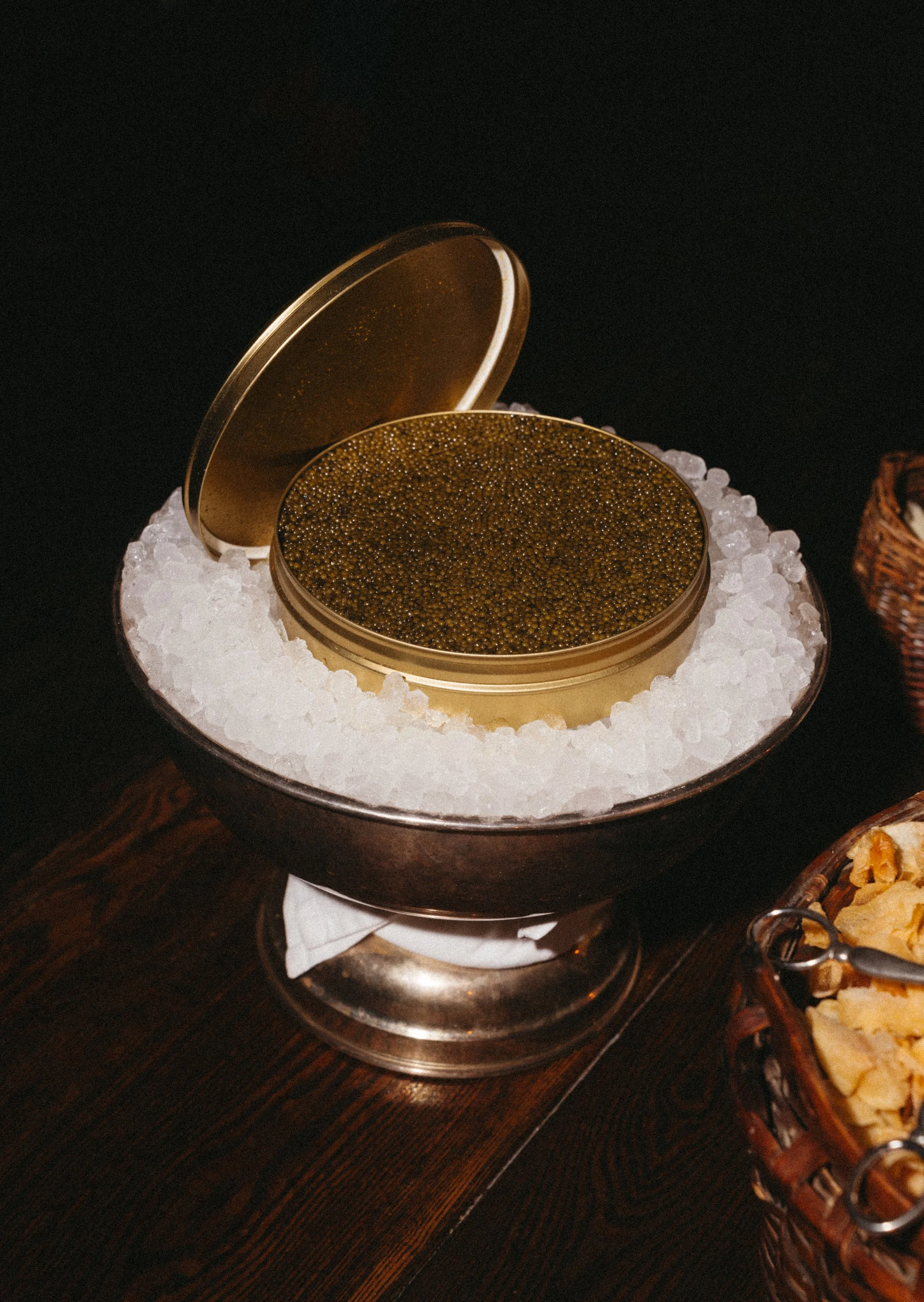A round, metal container with a hinged lid, filled with black caviar, sitting on a bed of ice in a bowl. The container's lid is slightly open.