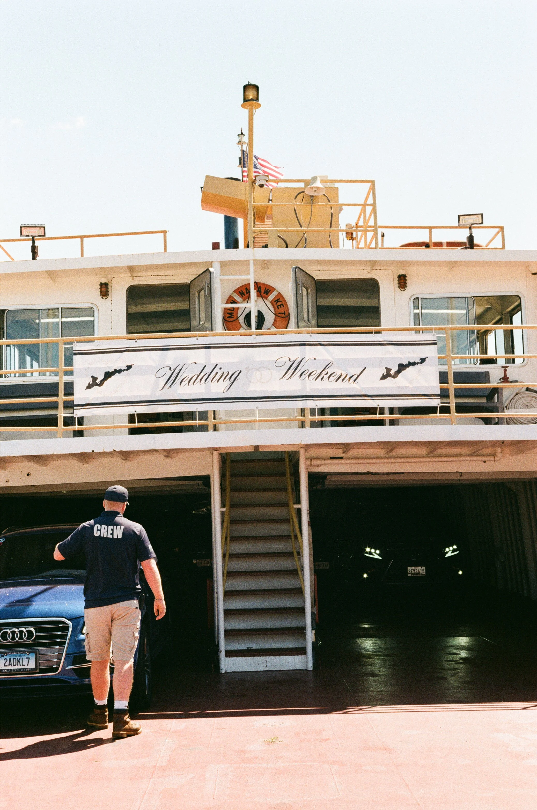 A ferry boat with a sign that reads 'Wedding Weekend' parked at a dock. A crew member is walking near parked cars underneath the ferry.